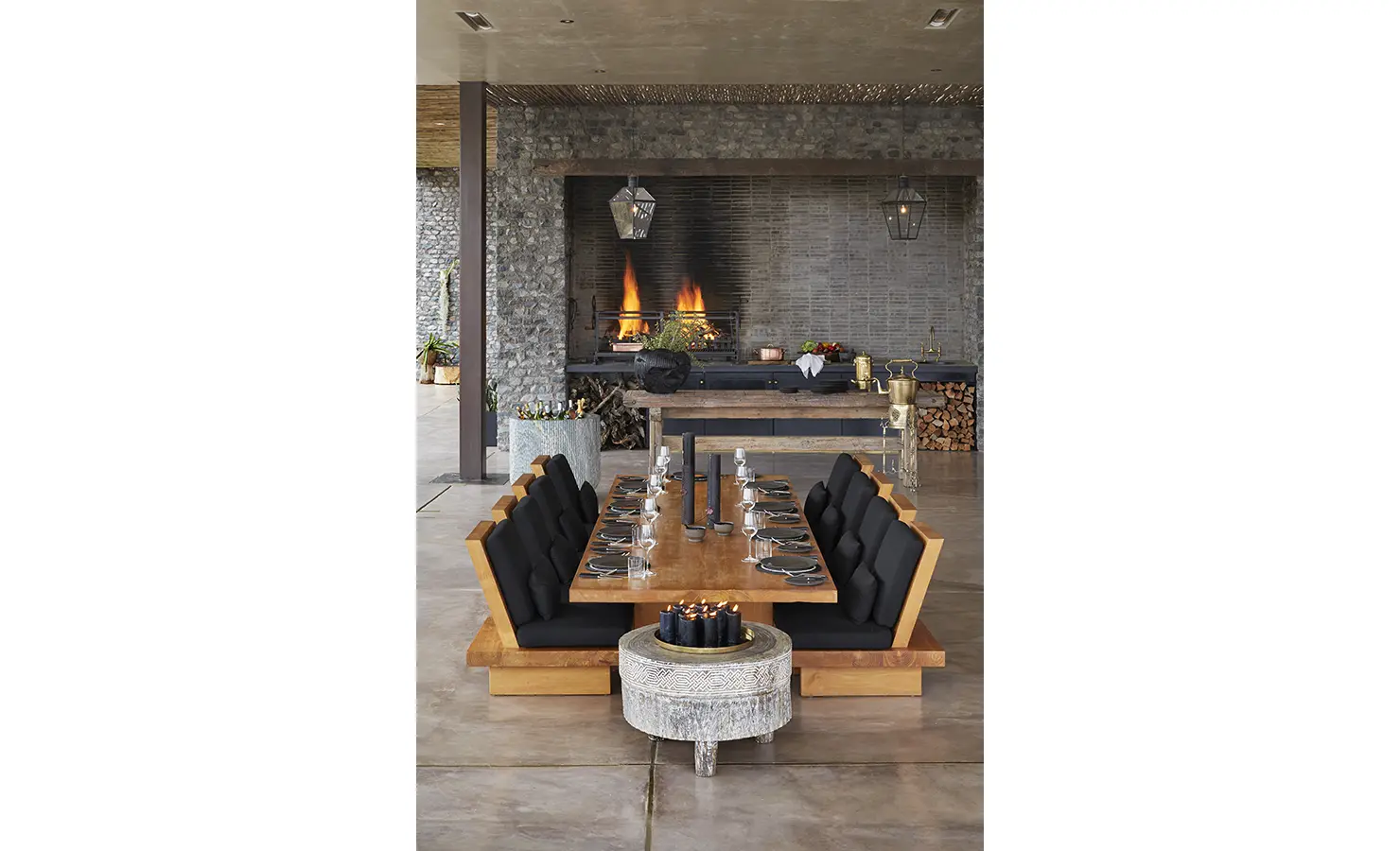 Perfect Hideaways: A stylish dining area featuring a long wooden table surrounded by modern chairs with black cushions. A large fireplace with a stone facade and decorative lanterns complete the inviting ambiance. Kubili House, Thornybush Game Reserve.