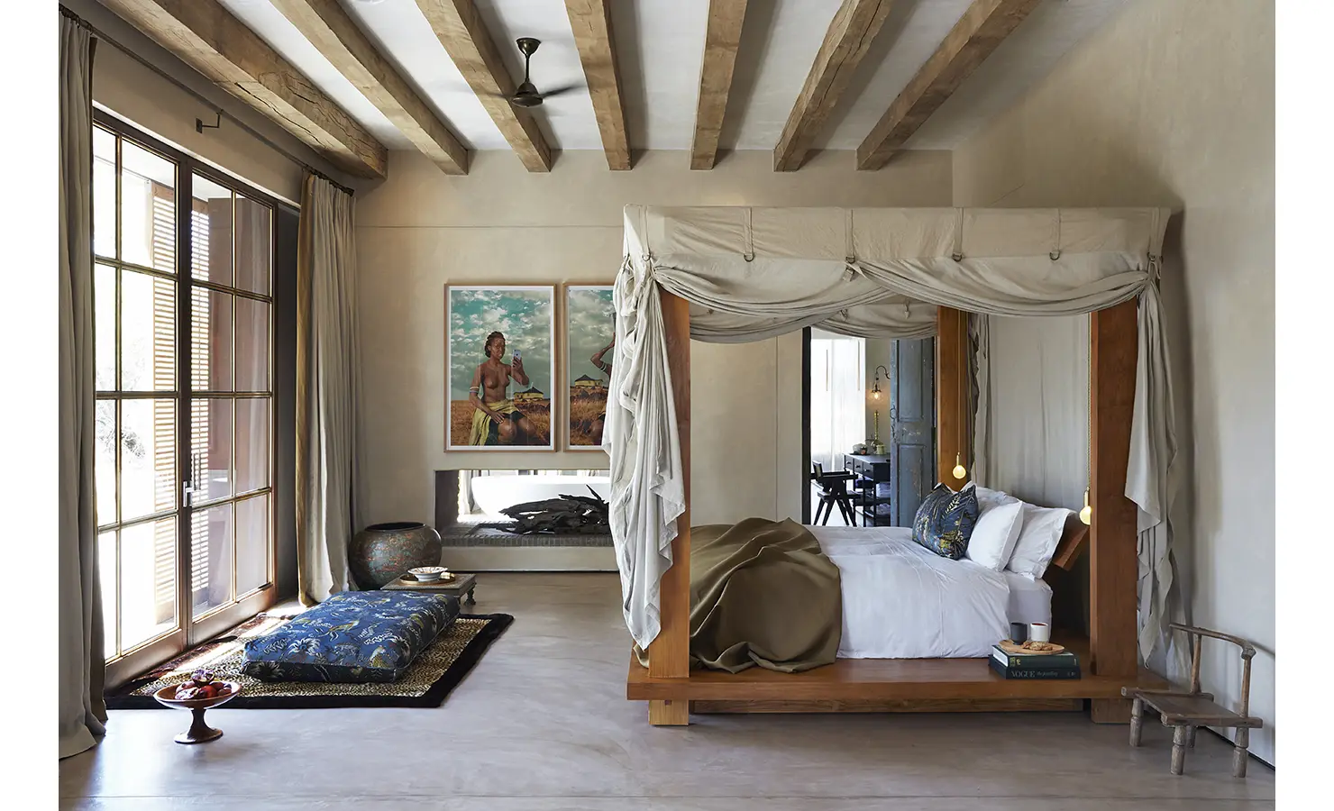 Perfect Hideaways: A bright bedroom with a canopy bed, exposed wooden beams, and a comfortable seating area near large windows draped with light curtains. Artistic wall decor adds a vibrant touch. Kubili House, Thornybush Game Reserve.
