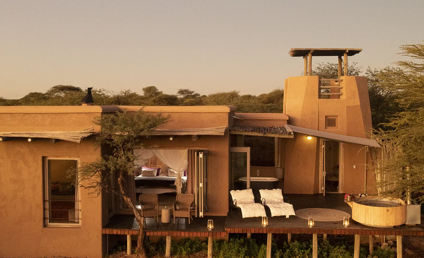 Perfect Hideaways: A luxurious suite with open-air verandas, a private outdoor bath, and an elevated viewing tower, surrounded by acacia trees and warm golden light. The Fort At Onguma, Etosha National Park.
