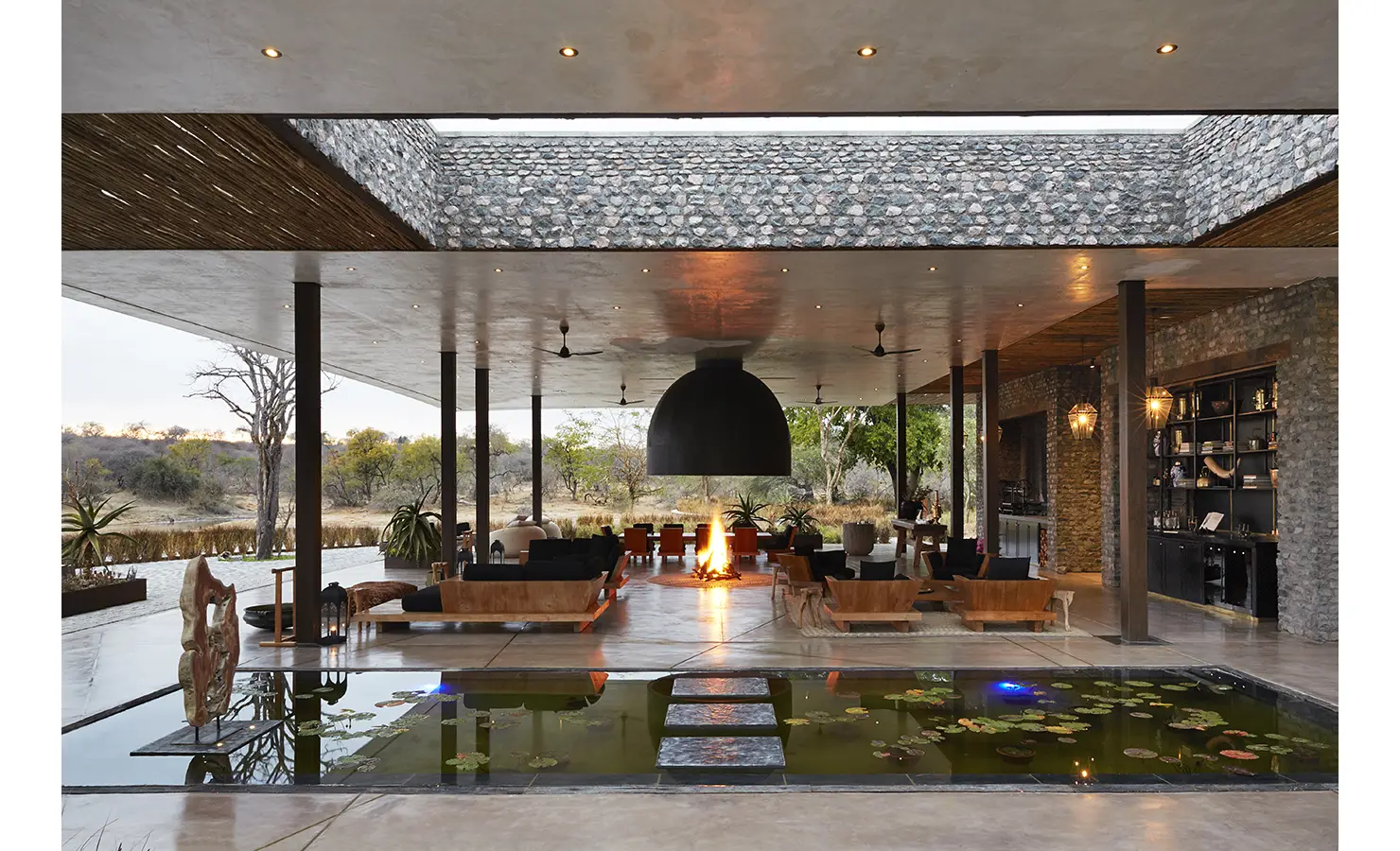 Perfect Hideaways: A stunning open-air living space with a fire pit at the centre, surrounded by modern furniture and overlooking a tranquil pond adorned with lily pads. Kubili House, Thornybush Game Reserve.