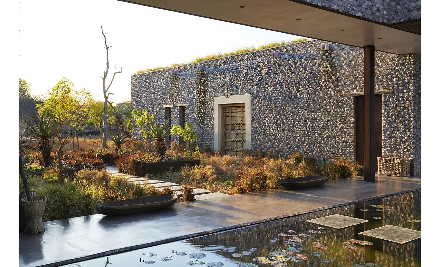 Perfect Hideaways: A lush garden with native plants and a pathway leading to a rustic stone building featuring a large wooden door. Kubili House, Thornybush Game Reserve.