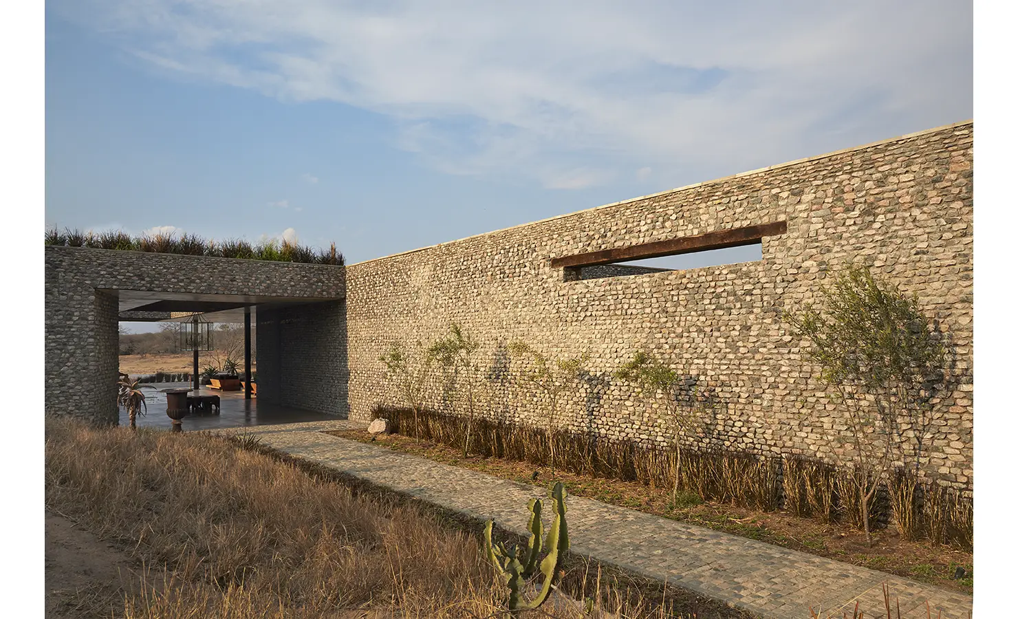 Perfect Hideaways: A long stone wall with clean lines and greenery along the path, opening to a sheltered outdoor seating area. Kubili House, Thornybush Game Reserve.
