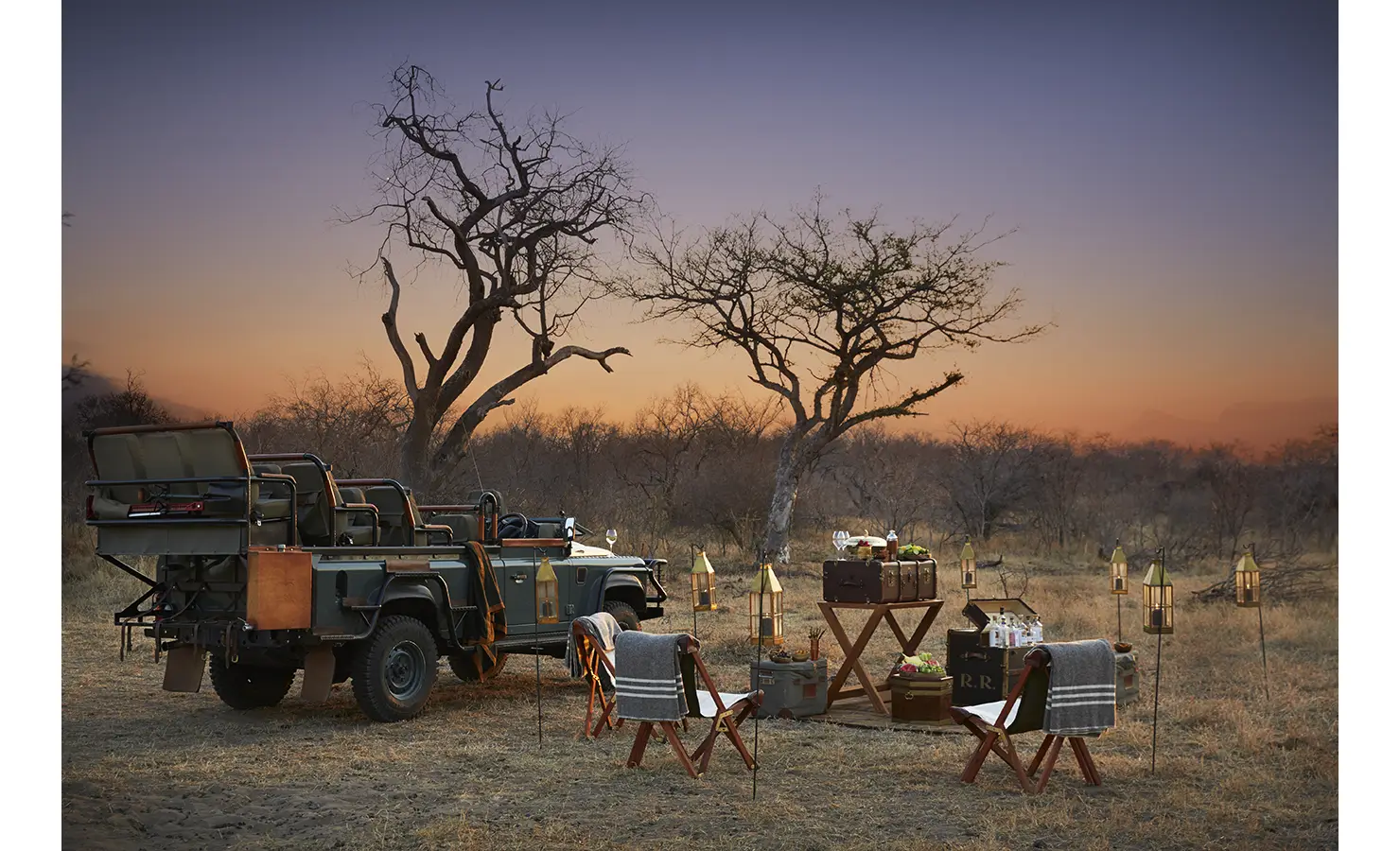 Perfect Hideaways: A bushveld dining setup at sunset, complete with a safari vehicle, folding chairs, lanterns, and a table set for an outdoor feast. Kubili House, Thornybush Game Reserve.