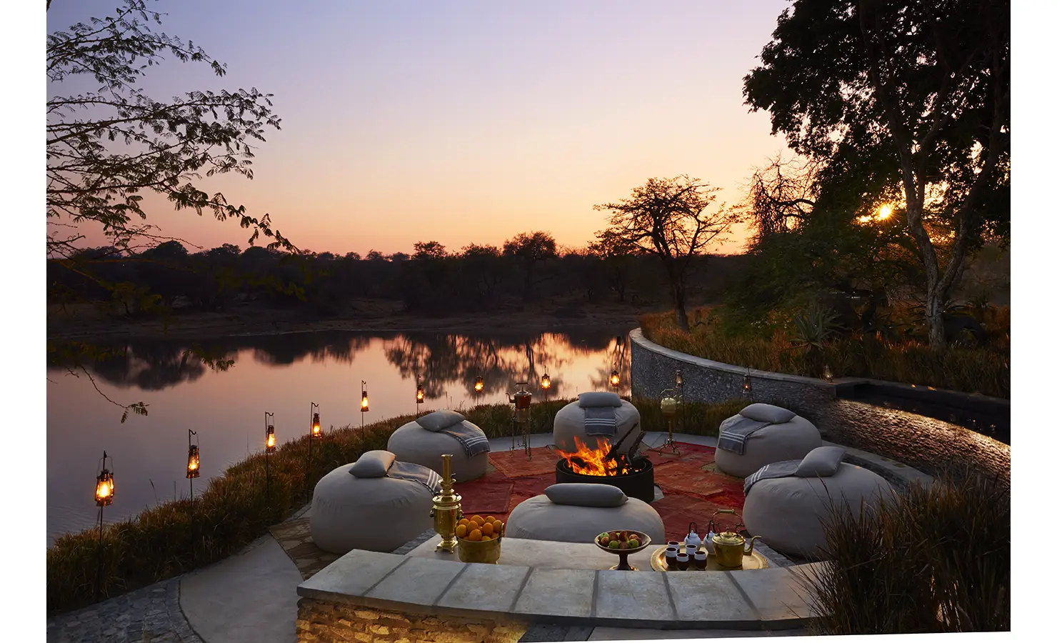 Perfect Hideaways: A serene firepit lounge set along a riverbank, surrounded by comfortable seating and illuminated by lanterns as the sun sets. Kubili House, Thornybush Game Reserve.