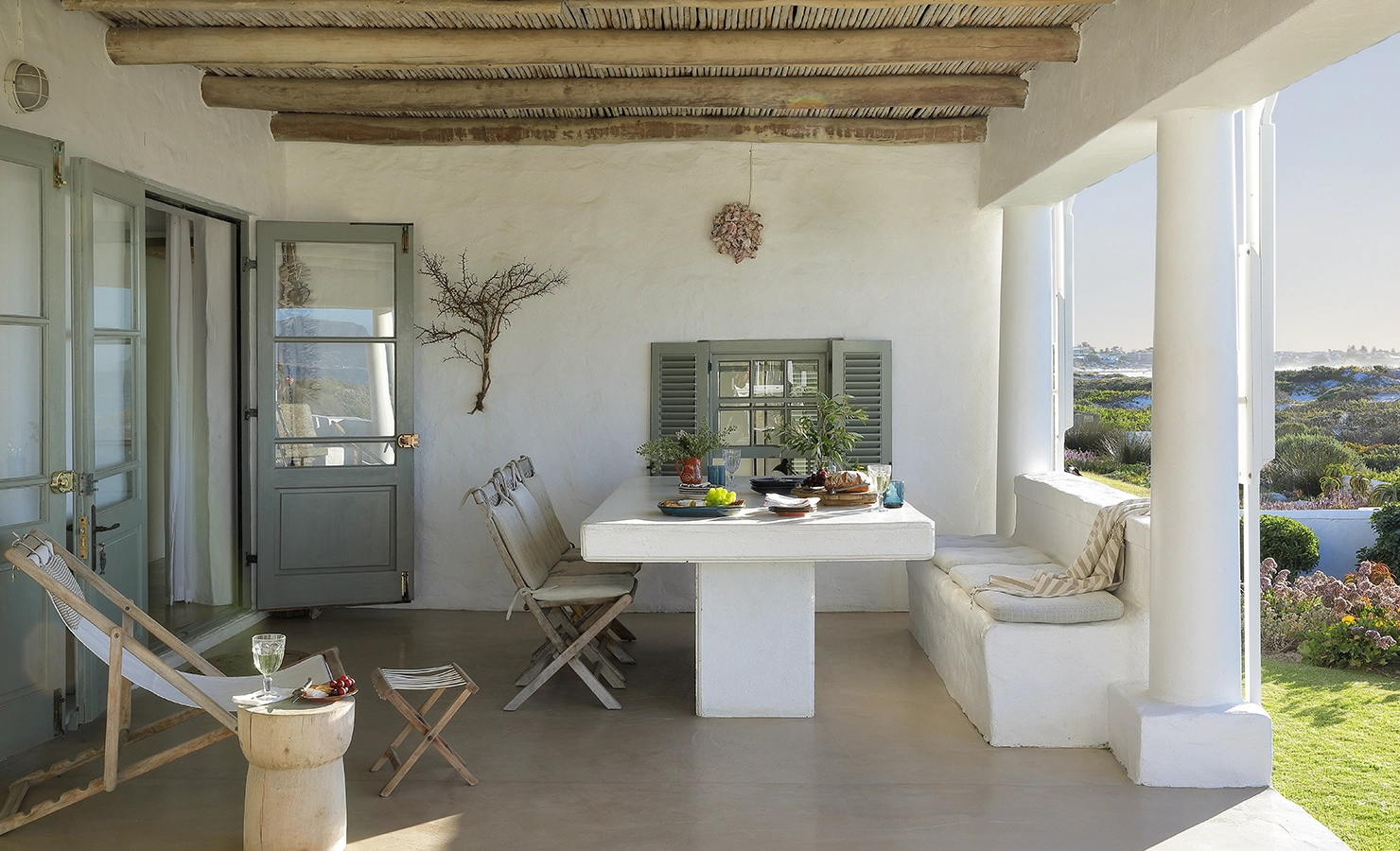 Perfect Hideaways: A spacious outdoor dining area on a veranda, featuring a white table, bench seating, and views of a lush garden and the ocean beyond. Farniente, Kommetjie.