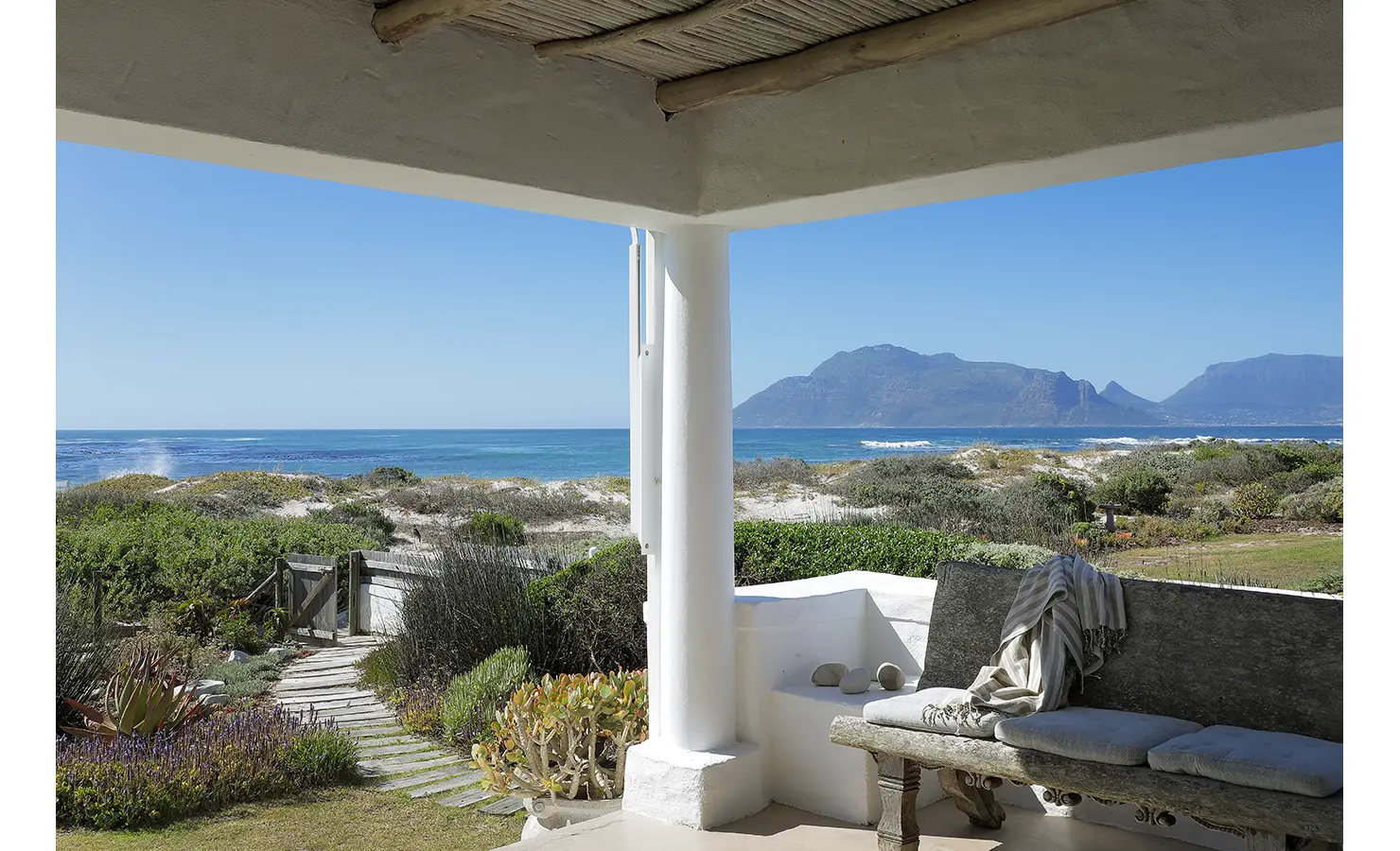 Perfect Hideaways: A serene view of the ocean framed by a rustic veranda with a wooden bench and soft cushions, overlooking a sandy pathway leading to the beach. Farniente, Kommetjie.