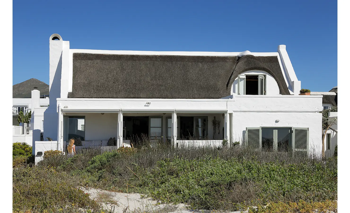 Perfect Hideaways: A whitewashed house with a thatched roof surrounded by coastal vegetation under a clear blue sky. Farniente, Kommetjie.