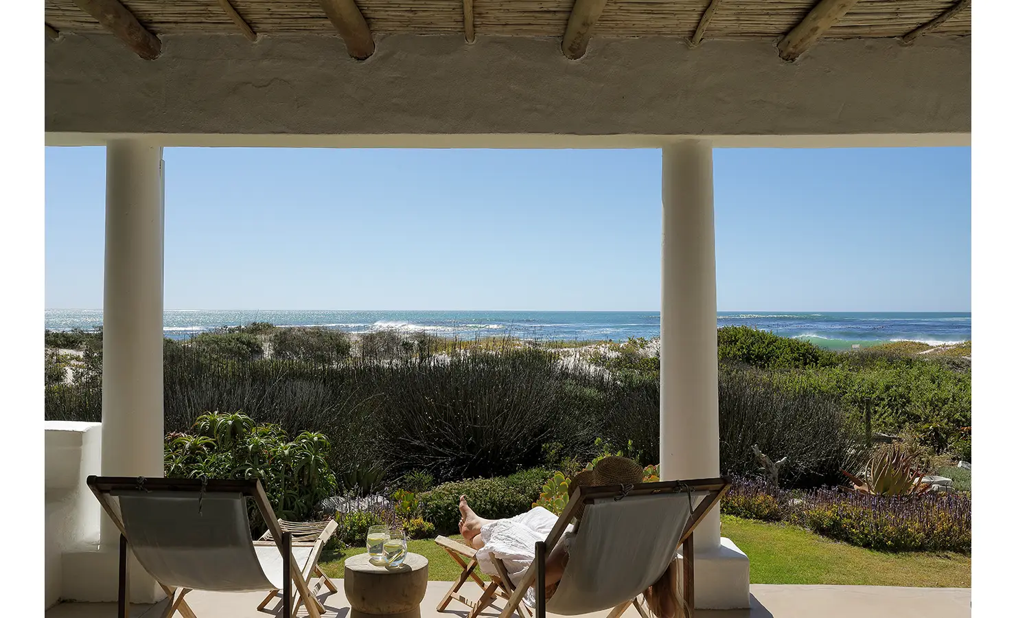 Perfect Hideaways: Two lounge chairs on a veranda facing the ocean, surrounded by lush greenery and dunes. A relaxing setting with a clear blue horizon. Farniente, Kommetjie.