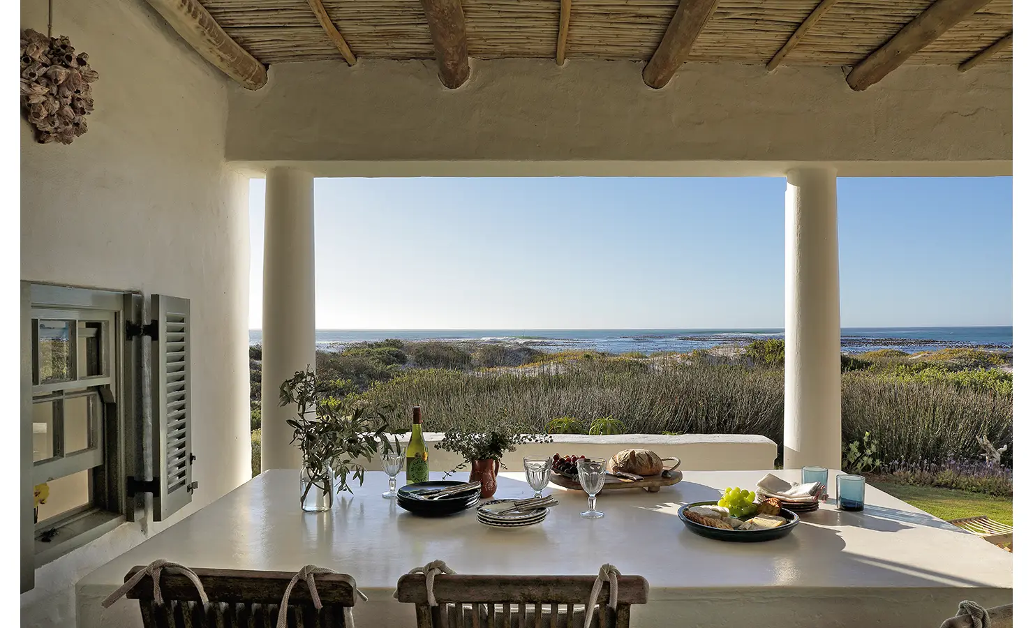 Perfect Hideaways: A covered outdoor dining area with a rustic table set for a meal, overlooking the ocean and coastal shrubs. Farniente, Kommetjie.