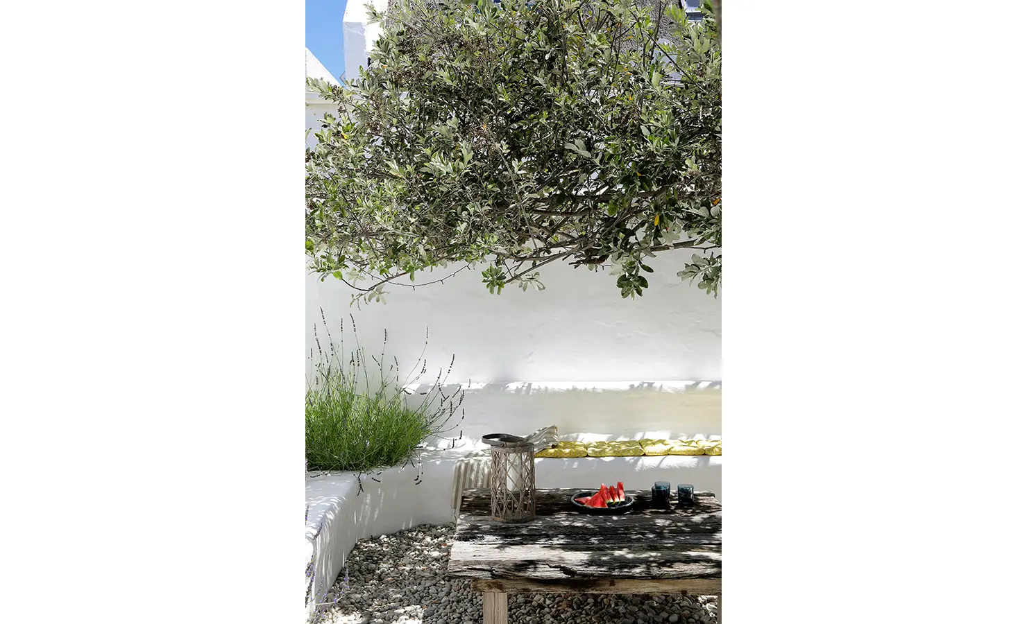 Perfect Hideaways: A shaded courtyard featuring a wooden table with watermelon slices, surrounded by pebbles, greenery, and a white wall. Farniente, Kommetjie.