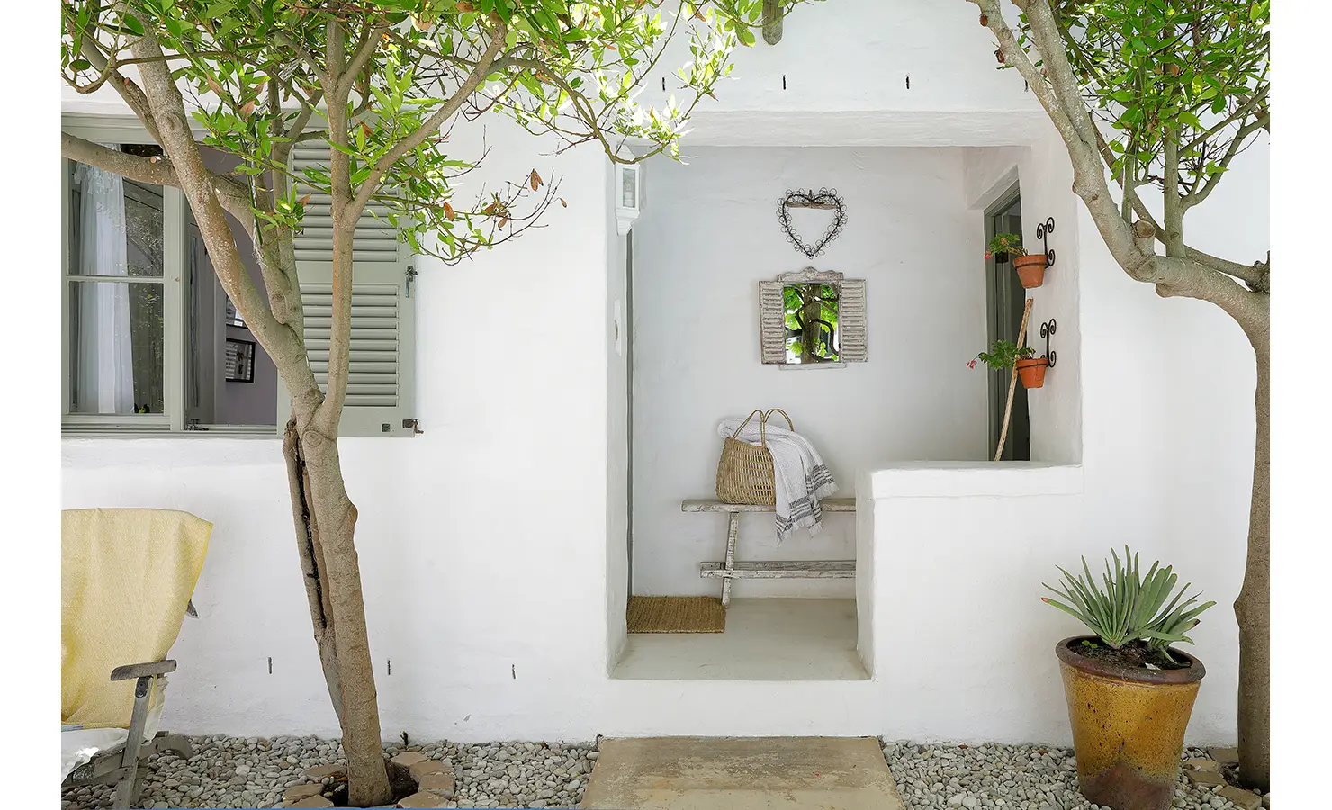 Perfect Hideaways: A charming whitewashed alcove with a bench adorned with a woven basket and a striped towel, surrounded by greenery and decorative elements. Farniente, Kommetjie.