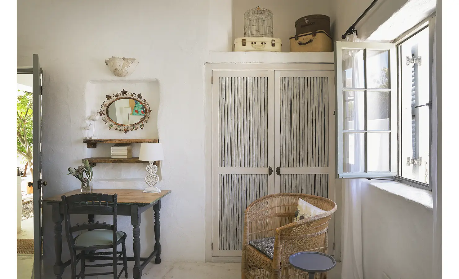 Perfect Hideaways: A cosy seating corner featuring a wicker chair, a writing desk, and a rustic cabinet with shutters, complemented by natural light streaming through an open window. Farniente, Kommetjie.