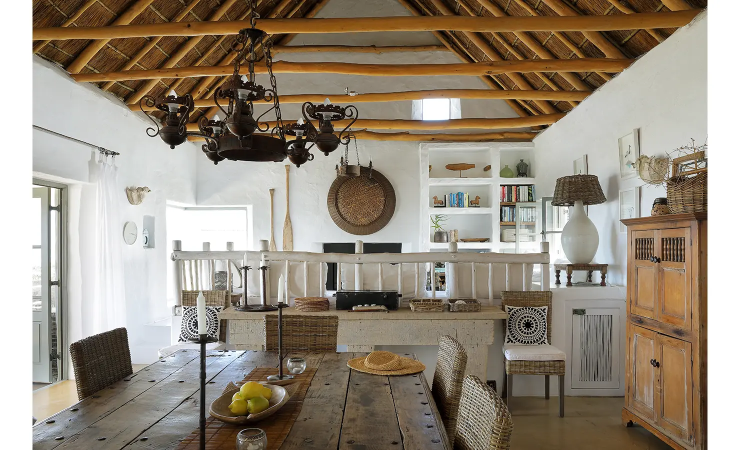 Perfect Hideaways: A stylish dining area with an expansive wooden table, wicker seating, and open shelving showcasing coastal decor under a high thatched ceiling. Farniente, Kommetjie.