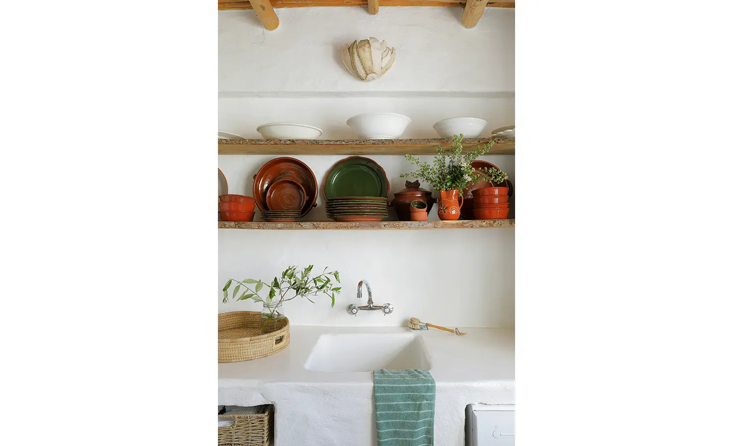 Perfect Hideaways: A rustic kitchen sink setup with open wooden shelves displaying vibrant terracotta and green dishes, complemented by a woven basket and a sprig of greenery. Farniente, Kommetjie.