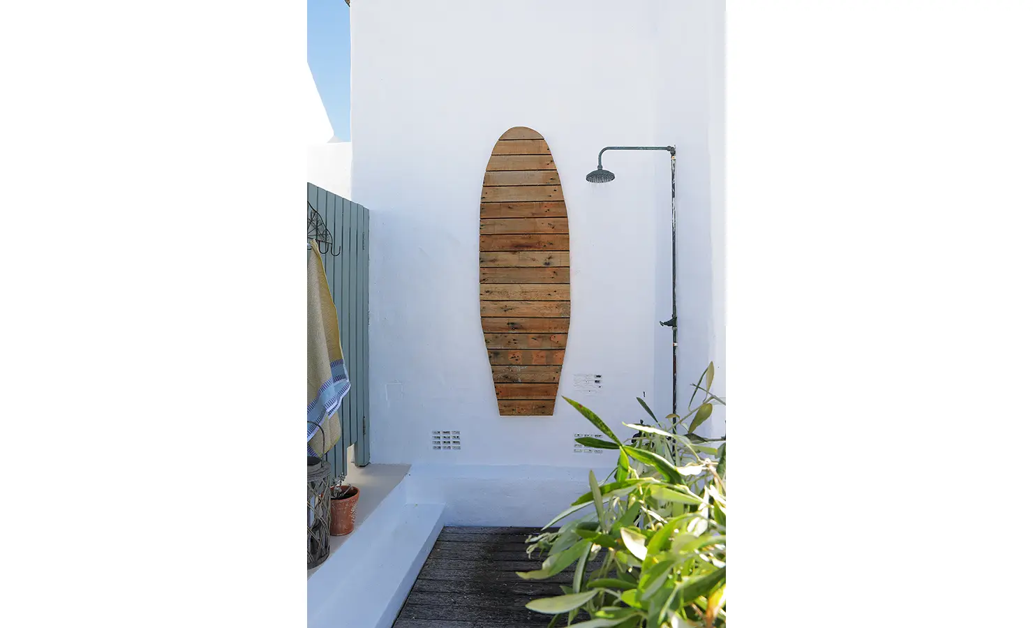 Perfect Hideaways: An outdoor shower area with a surfboard-shaped wooden panel on a white wall, surrounded by coastal greenery. Farniente, Kommetjie.
