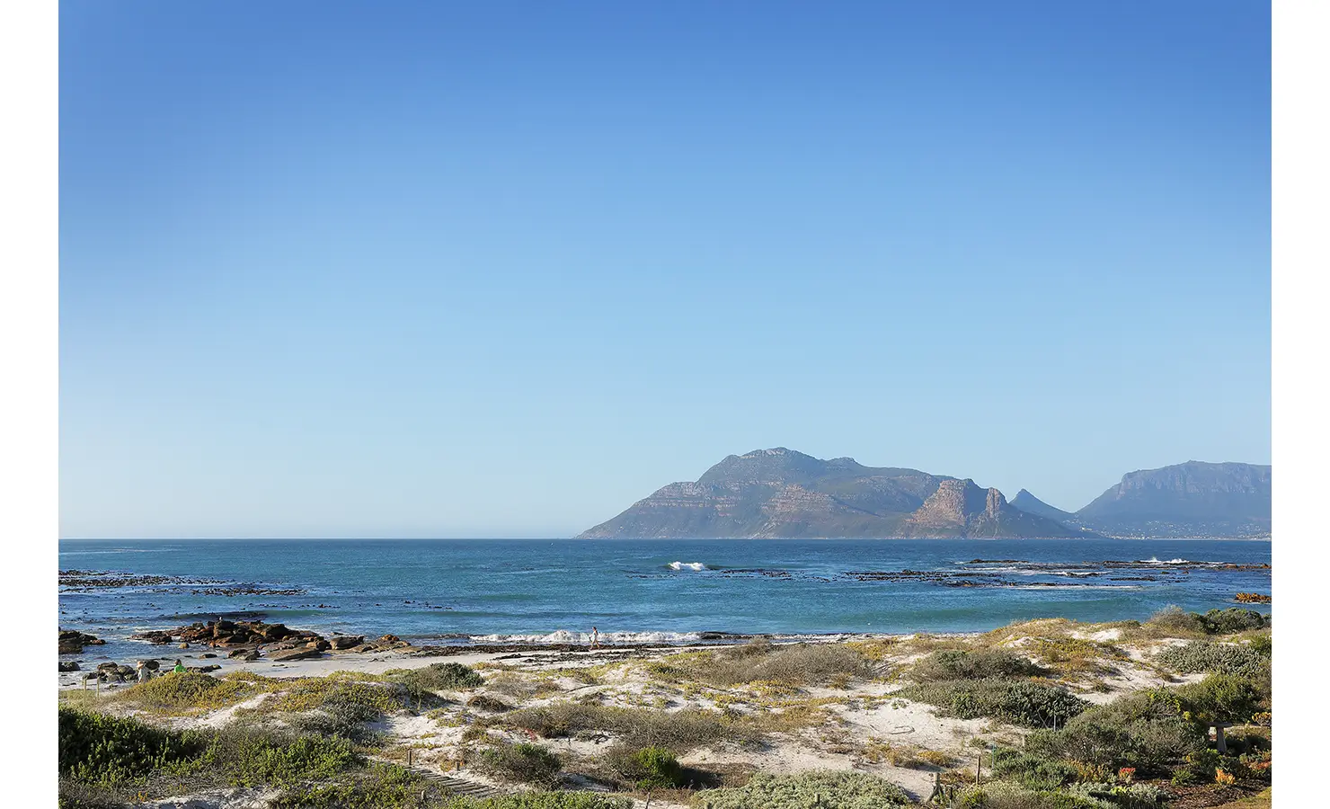 Perfect Hideaways: A pristine view of the ocean and mountains, framed by sandy dunes and coastal vegetation under a clear blue sky. Farniente, Kommetjie.