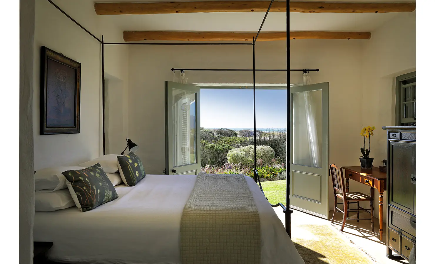 Perfect Hideaways: A bright and airy bedroom with double doors opening to a lush garden, featuring a cosy bed with botanical-themed cushions and a wooden desk. Farniente, Kommetjie.
