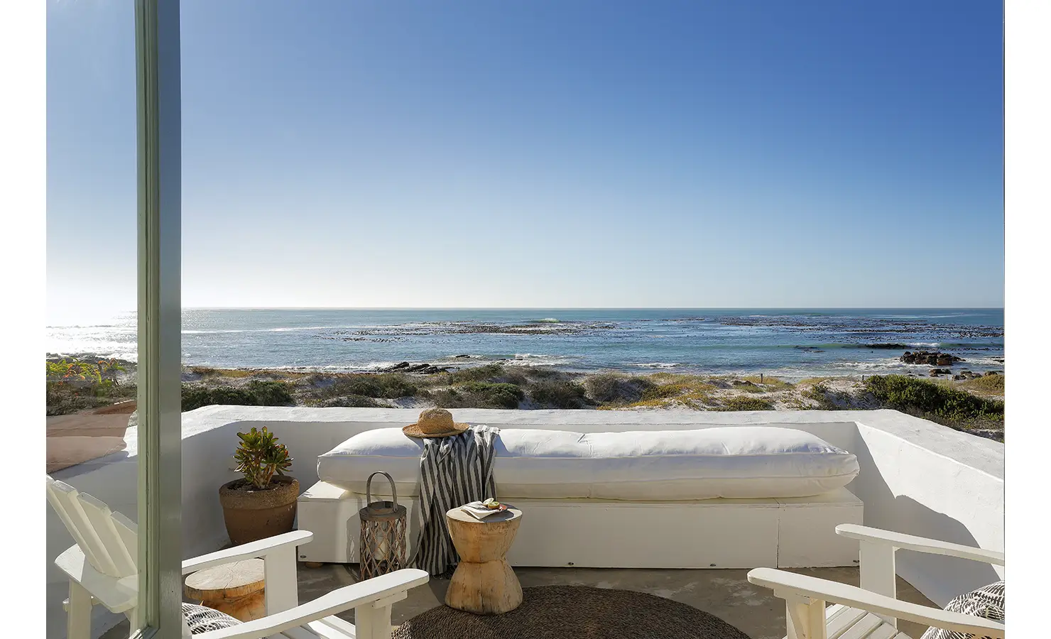 Perfect Hideaways: A serene terrace overlooking the ocean, with white seating, wooden stools, and a straw hat resting on a cushion. Farniente, Kommetjie.