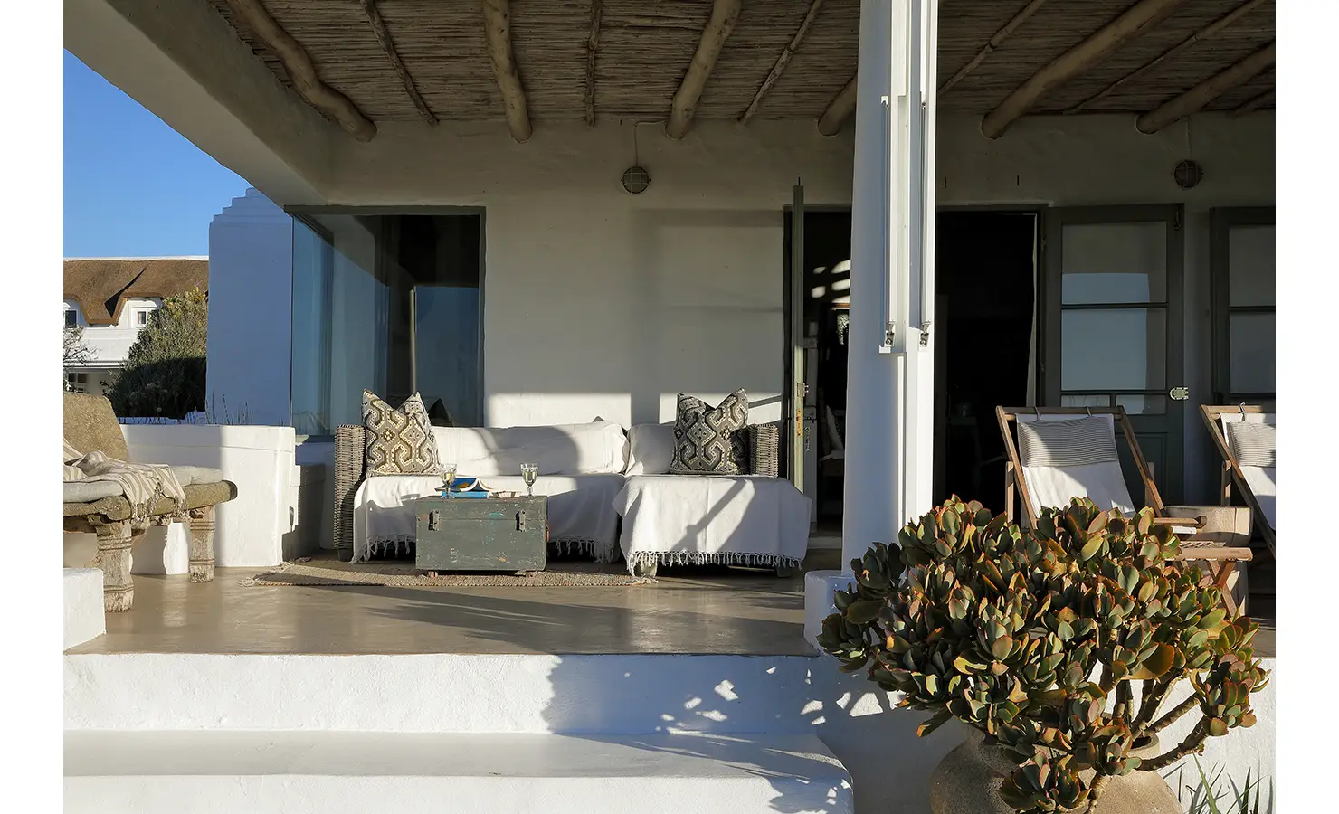 Perfect Hideaways: A covered veranda with cushioned seating and a wooden table, styled with coastal decor and bathed in natural light. Farniente, Kommetjie.