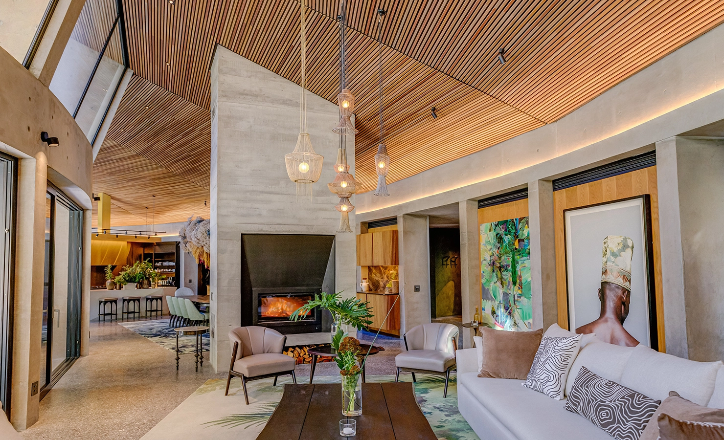 Perfect Hideaways: A striking modern living space featuring a soaring timber-lined ceiling, concrete walls, and an elegant fireplace, with curated contemporary artwork, soft neutral-toned furnishings, and pendant lighting adding warmth and texture to the sophisticated interior – a seamless blend of architectural precision and inviting comfort. Villa Verte, Hout Bay.