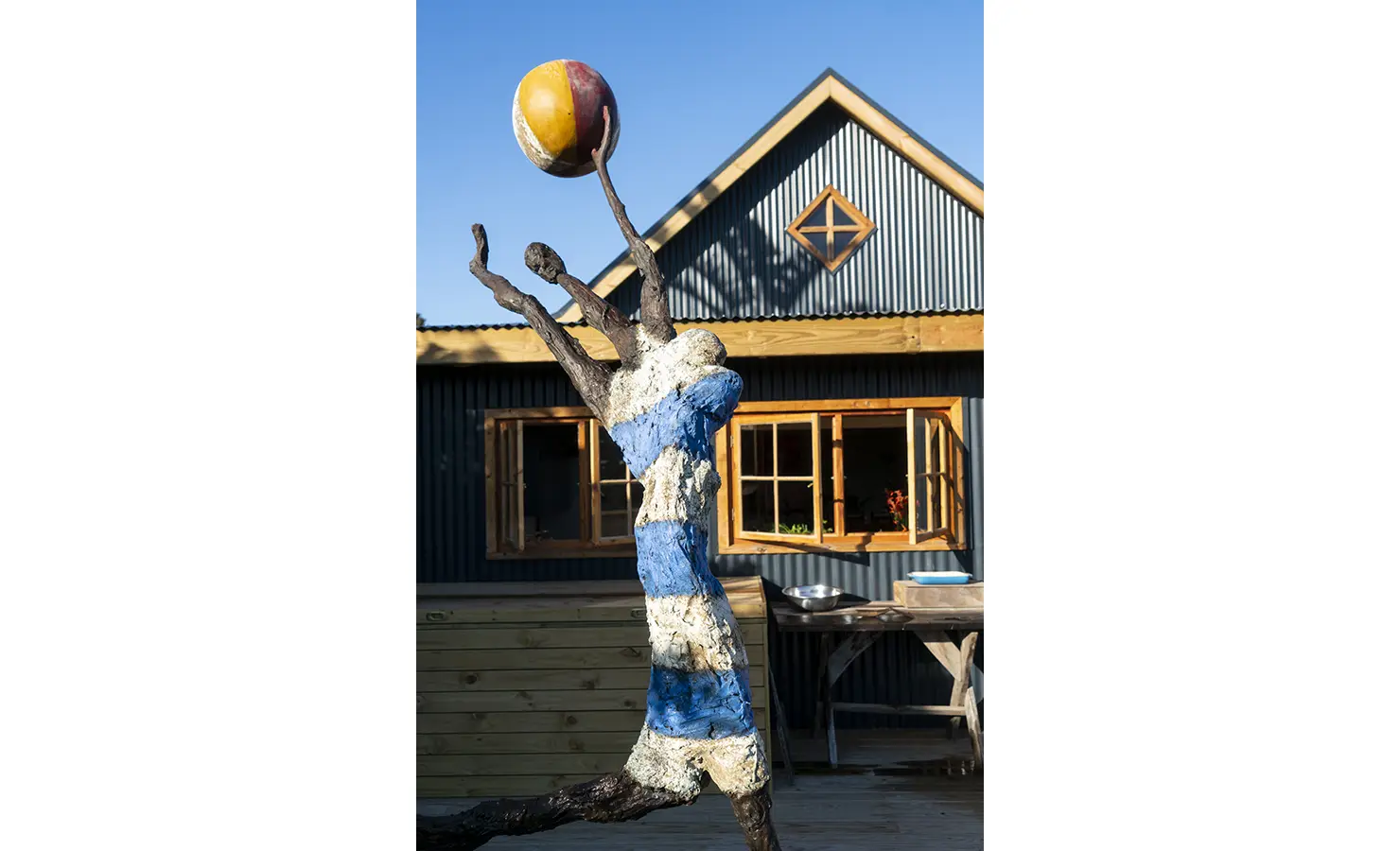 Perfect Hideaways: A creative sculpture of a figure with outstretched arms holding a colourful ball, set against a corrugated house exterior with open wooden windows. Bird House, Plettenberg Bay.
