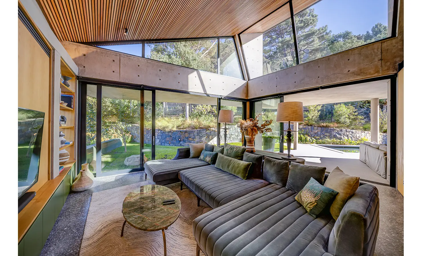 Perfect Hideaways: A sun-filled lounge with deep green velvet seating, panoramic glass walls, and views of the lush garden, merging indoor comfort with natural serenity. Villa Verte, Hout Bay.