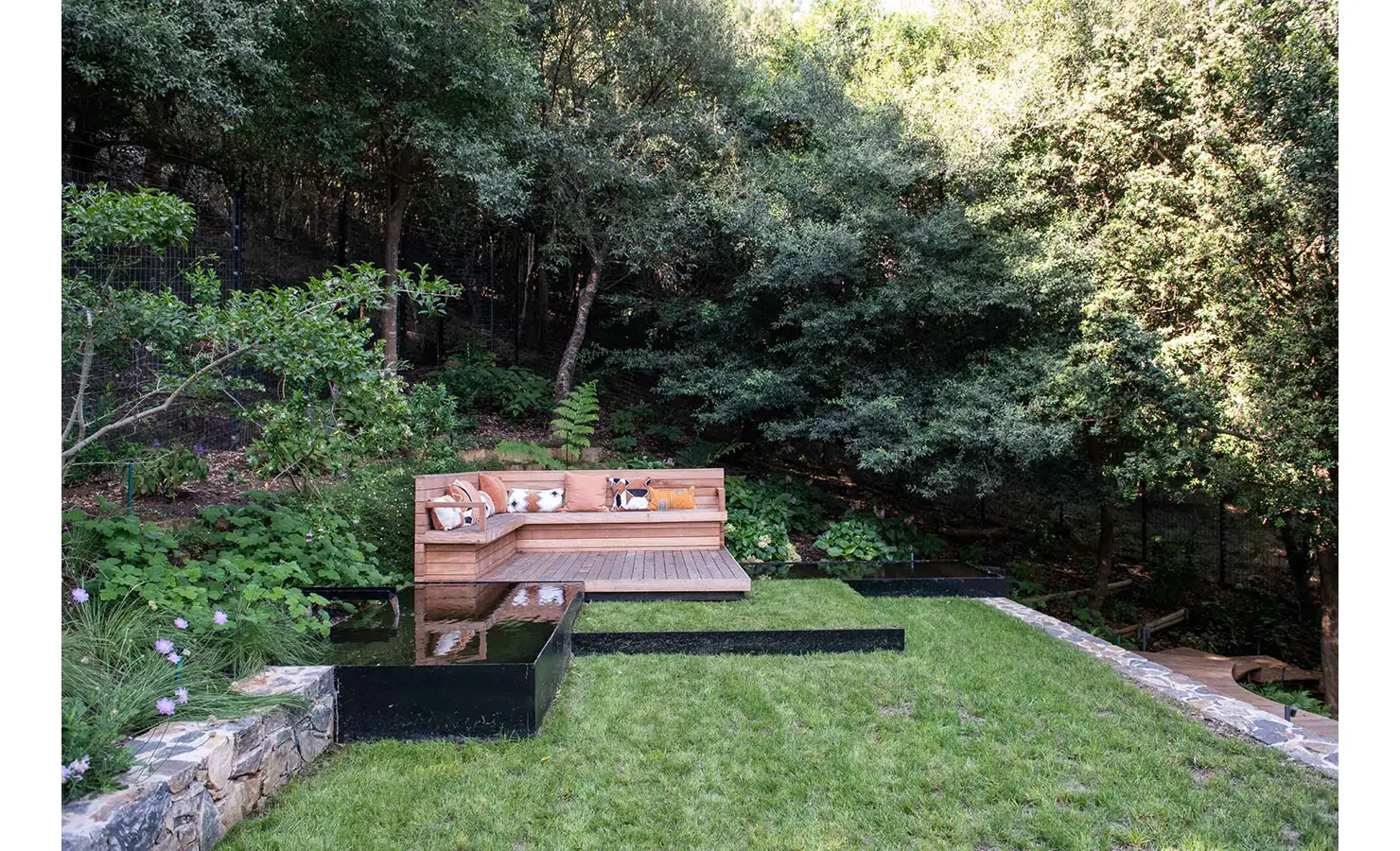 Perfect Hideaways: A tranquil garden nook featuring a custom timber bench with vibrant cushions, surrounded by dense forest and reflective water features, perfect for peaceful retreat. Villa Verte, Hout Bay.
