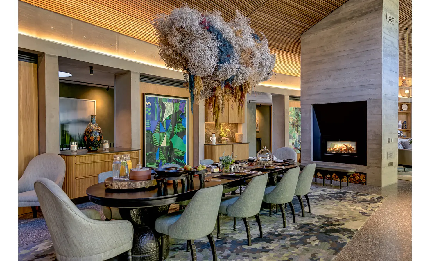 Perfect Hideaways: A dramatic dining space with soft sage chairs, a bold hanging floral installation, colourful abstract artwork, and a sleek double-sided fireplace, offering a theatrical yet welcoming setting. Villa Verte, Hout Bay.