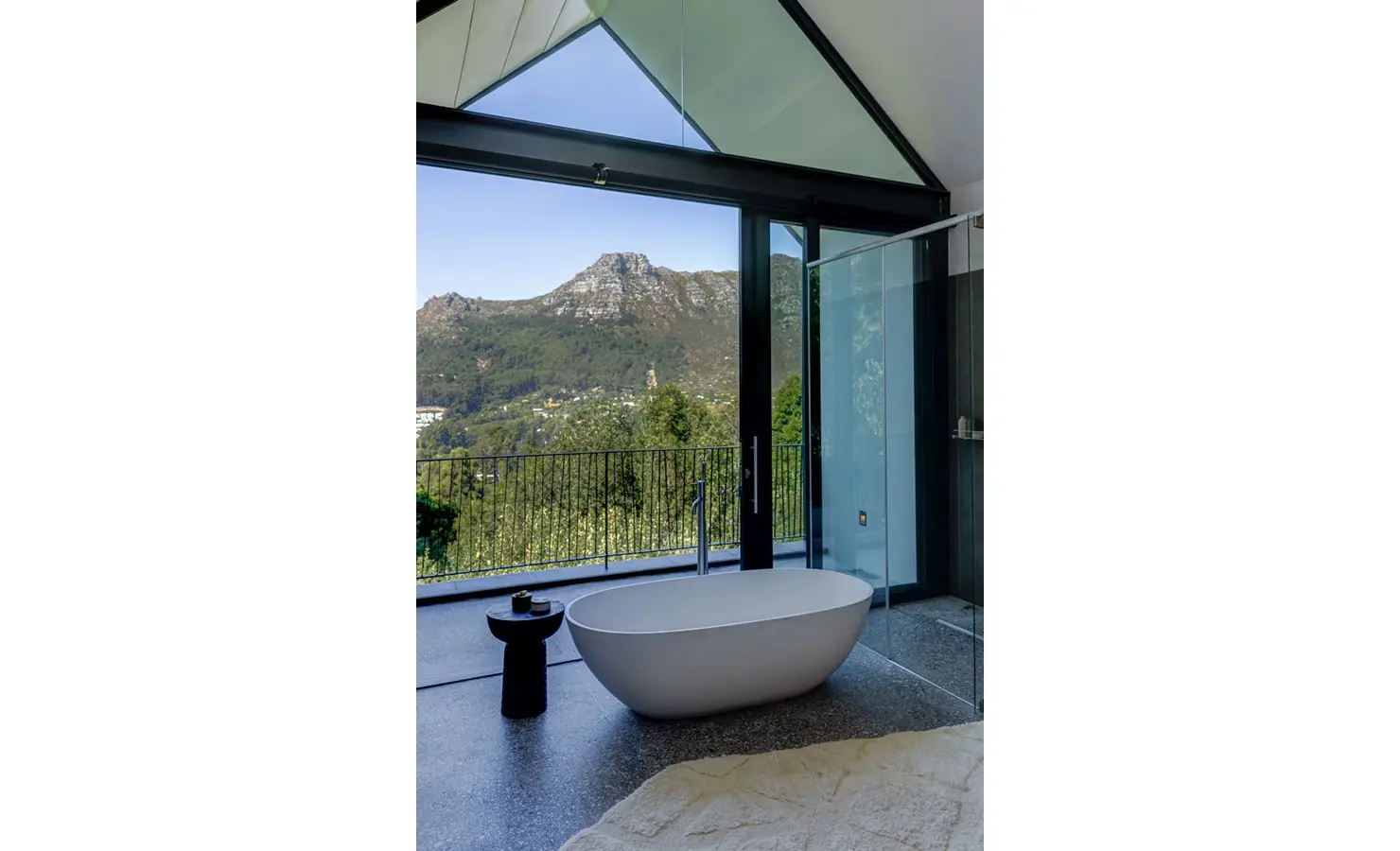 Perfect Hideaways: A serene bathroom retreat with a freestanding tub positioned before full-height glass doors, offering panoramic mountain views framed by a vaulted ceiling. Villa Verte, Hout Bay.