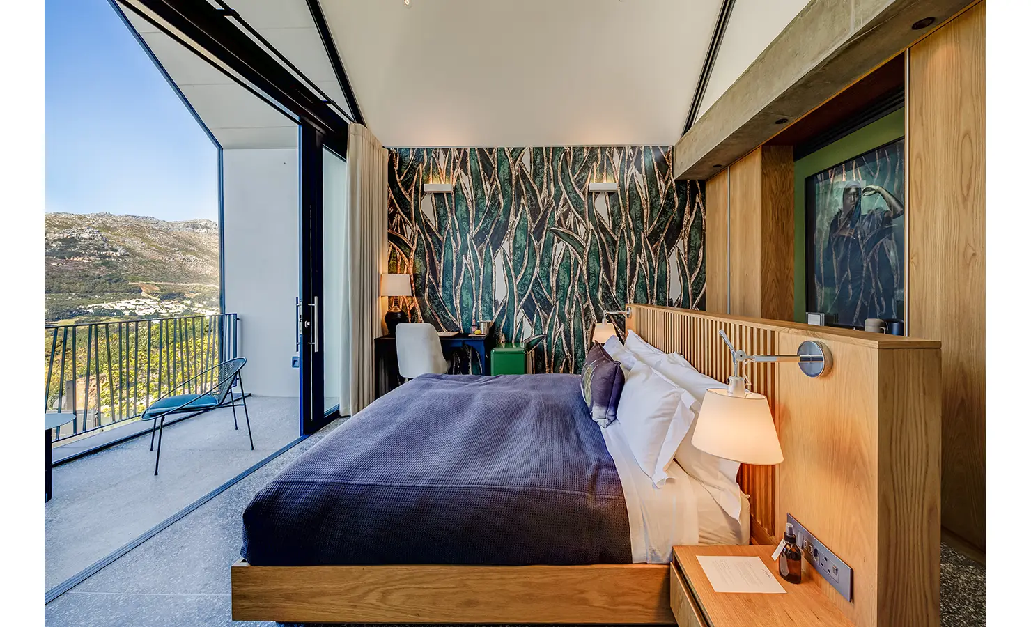 Perfect Hideaways: A bold bedroom with dramatic leafy wallpaper, minimalist furnishings, and a large balcony offering uninterrupted mountain vistas and fresh air. Villa Verte, Hout Bay.