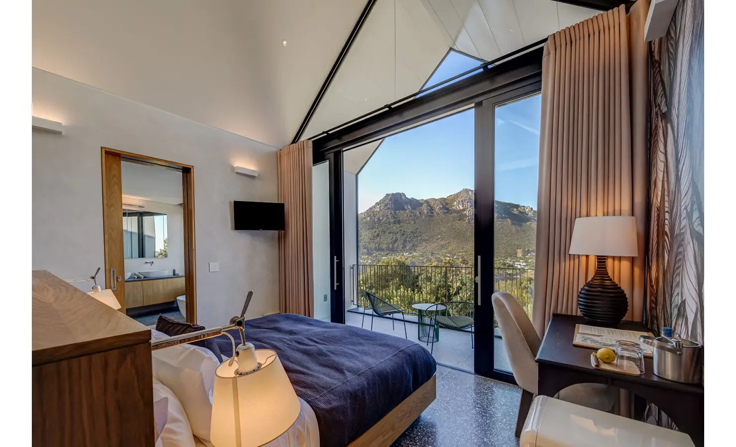 Perfect Hideaways: A modern bedroom with soft lighting and desk space, opening onto a mountain-facing balcony through tall glass doors, inviting morning sunlight and peaceful views. Villa Verte, Hout Bay.