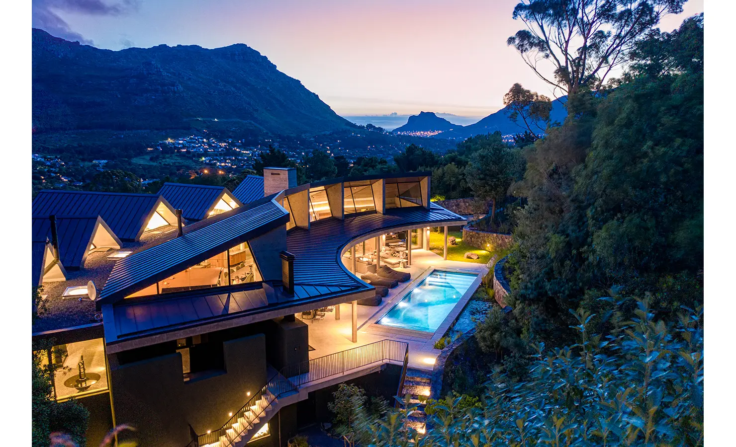 Perfect Hideaways: A magical twilight scene of the villa aglow with warm lights, its curved architecture embracing a lit pool and forested hillside, with distant views of the sea and mountains beyond. Villa Verte, Hout Bay.
