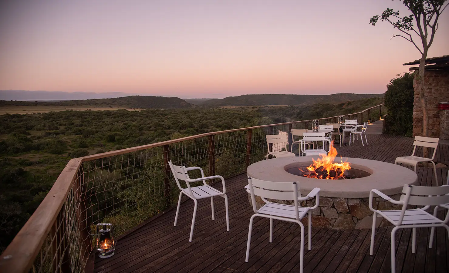 Perfect Hideaways: A deck with a central firepit, surrounded by chairs and offering stunning views of the lush green landscape at dusk. Riverdene, Shamwari Private Game Reserve.