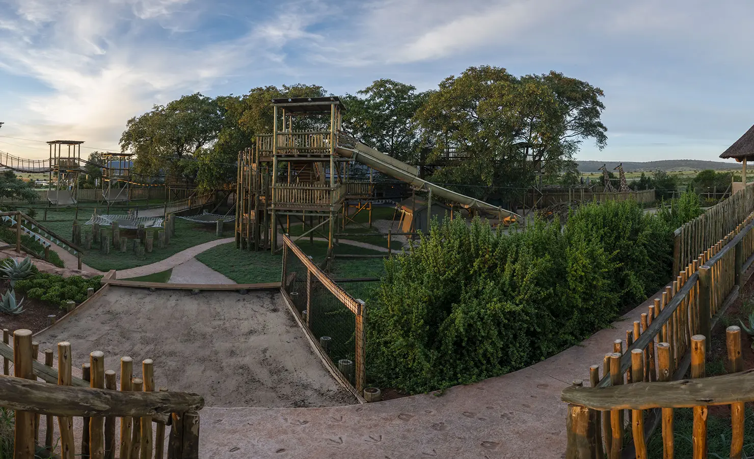 Perfect Hideaways: An expansive outdoor play area with wooden climbing structures, slides, and greenery, set under a blue sky. Riverdene, Shamwari Private Game Reserve.