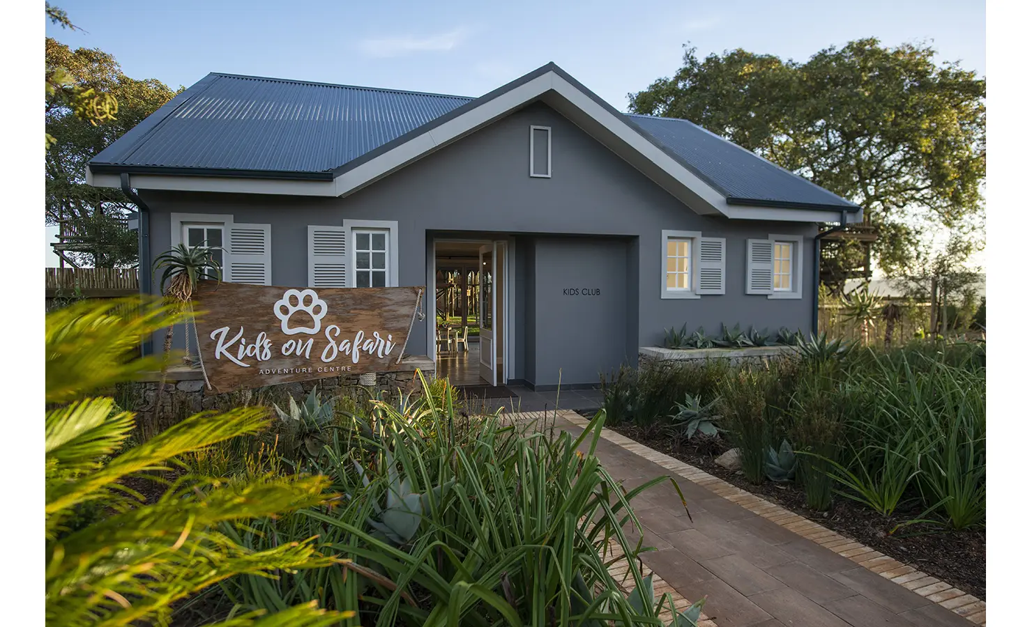 Perfect Hideaways: A charming grey kids’ club building surrounded by lush greenery, with a sign reading "Kids on Safari Adventure Centre." Riverdene, Shamwari Private Game Reserve.