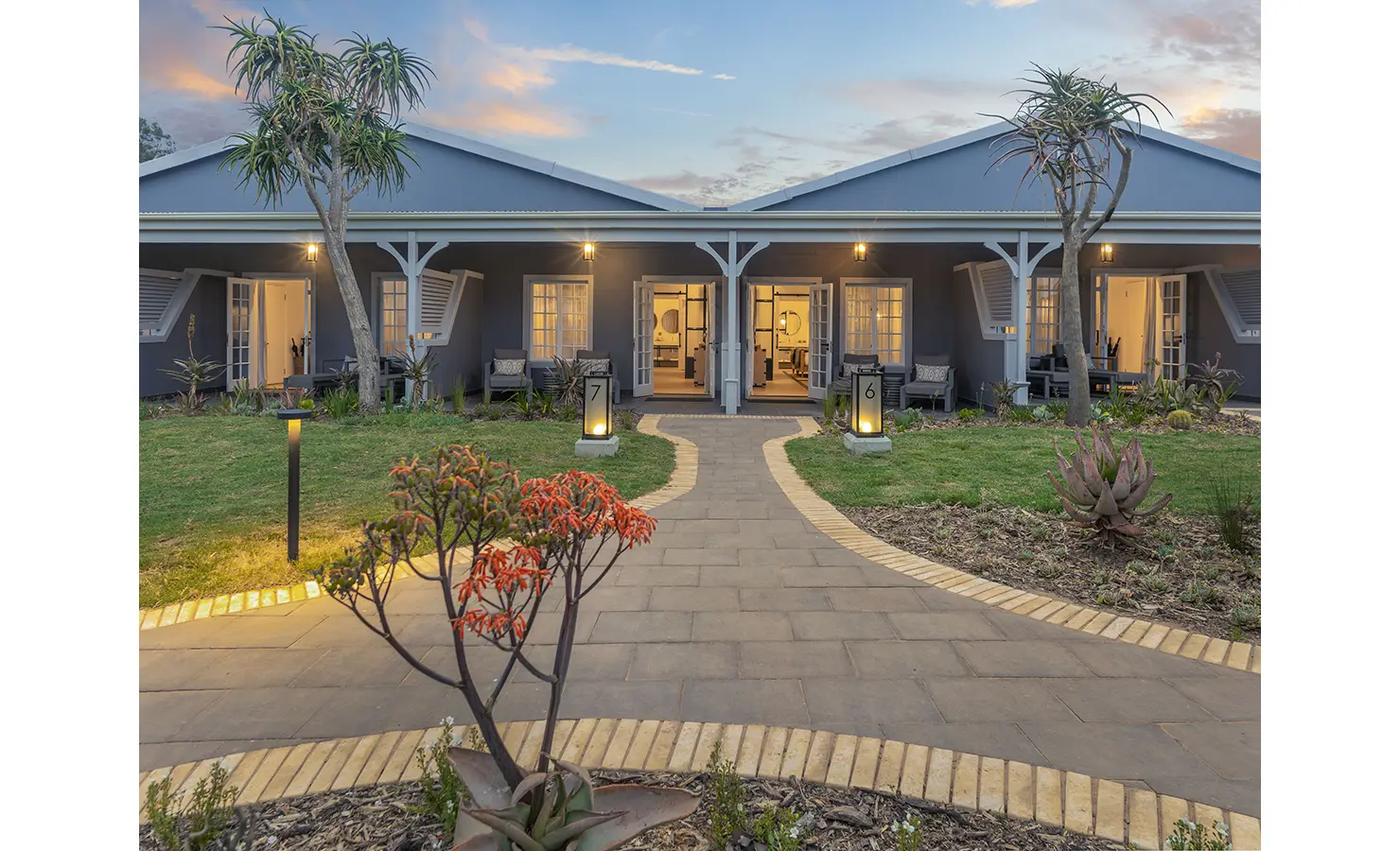 Perfect Hideaways: A beautifully landscaped pathway leading to two elegant lodge suites with soft exterior lighting during sunset. Riverdene, Shamwari Private Game Reserve.