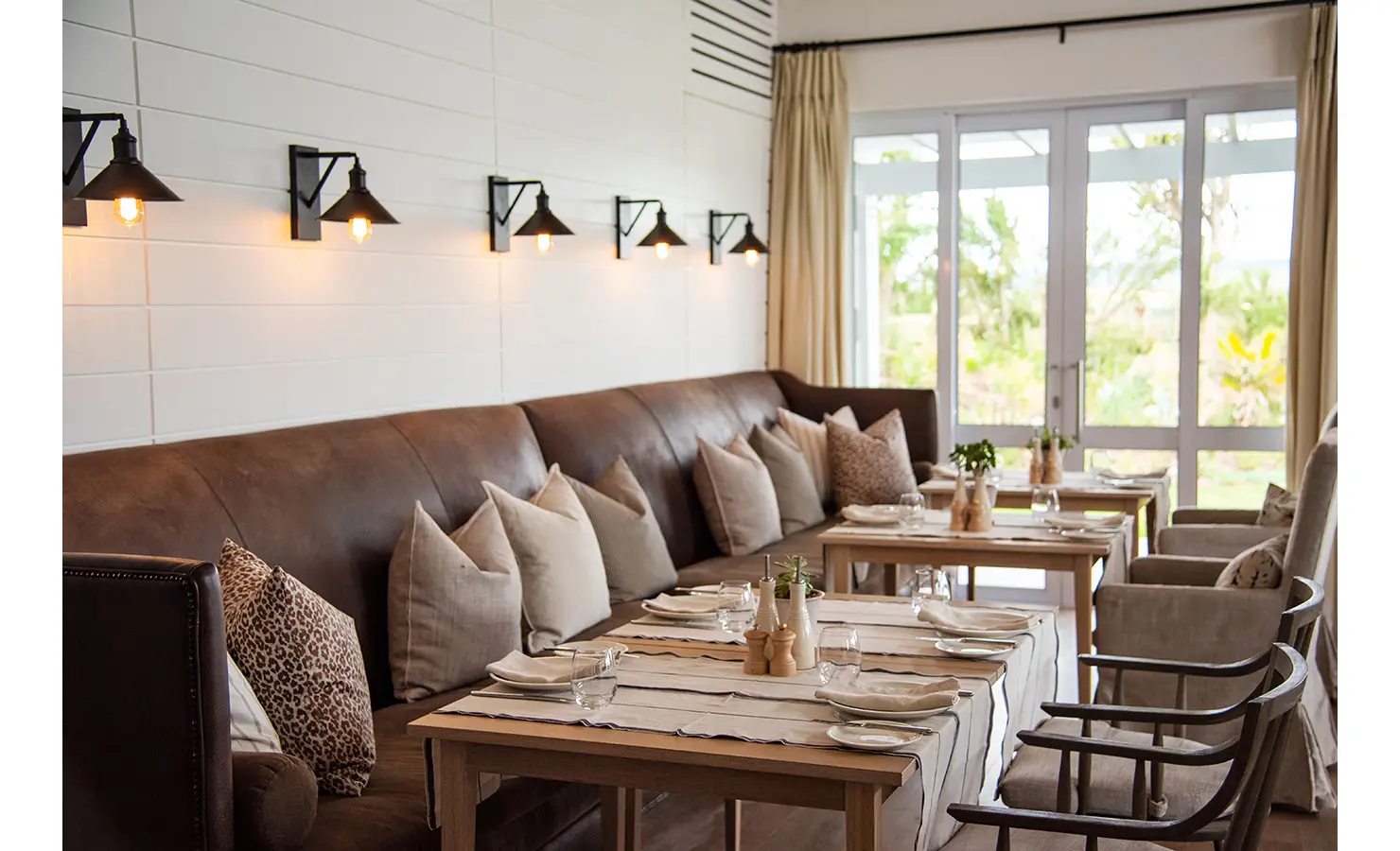 Perfect Hideaways: A cosy dining area with leather seating, wooden tables set for meals, and views of a lush garden through glass doors. Riverdene, Shamwari Private Game Reserve.