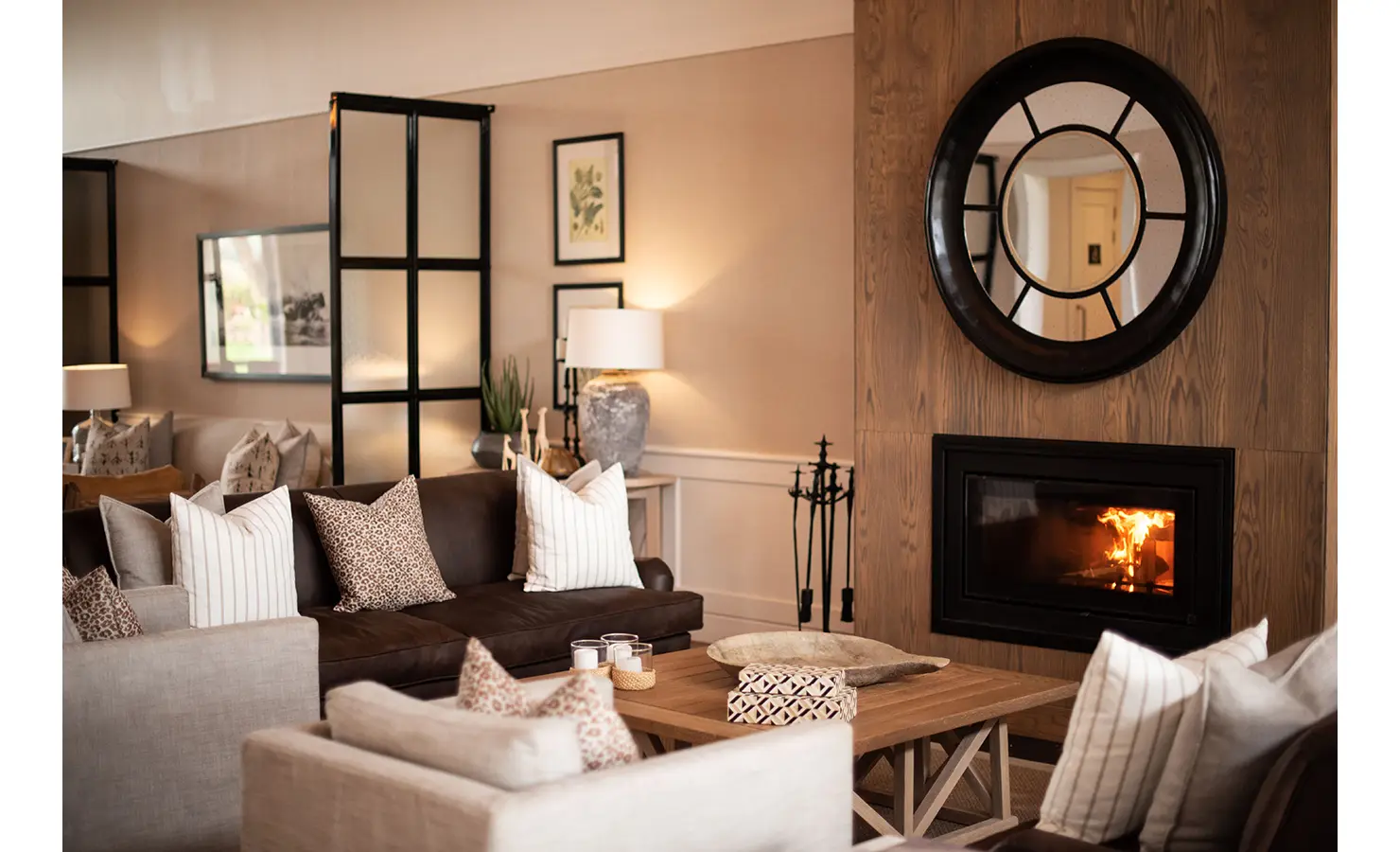 Perfect Hideaways: A cosy lounge area featuring a fireplace with a circular mirror above it, a brown leather sofa, and neutral-toned cushions. Riverdene, Shamwari Private Game Reserve.