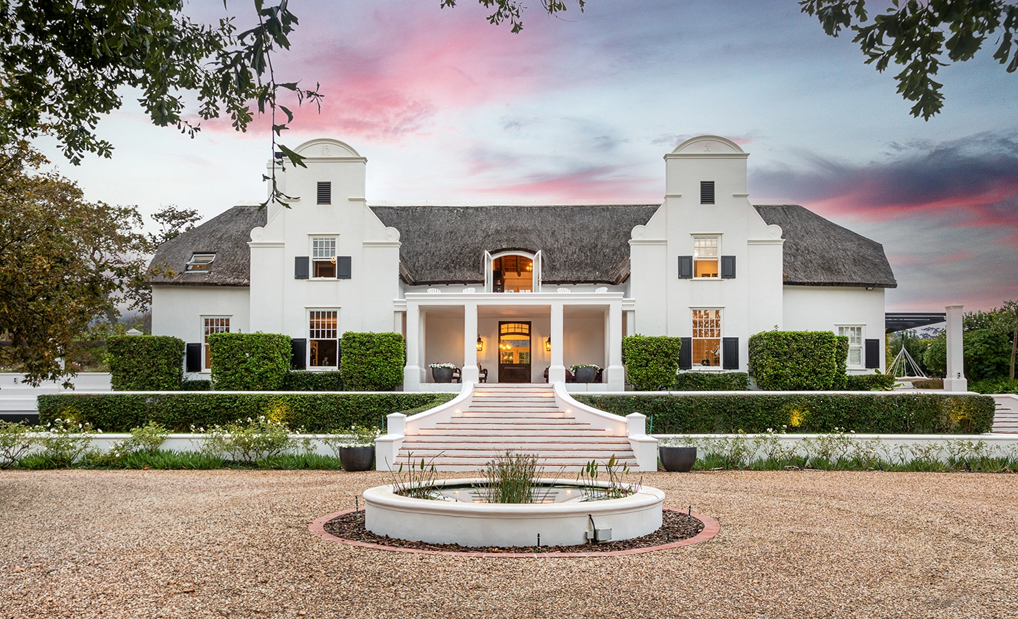 Perfect Hideaways: A grand Cape Dutch manor with symmetrical gables, a thatched roof, and elegant entrance steps flanked by manicured hedges and a central fountain, set against a dramatic pink and blue sunset sky — Buitenzorg Manor House, Constantia.