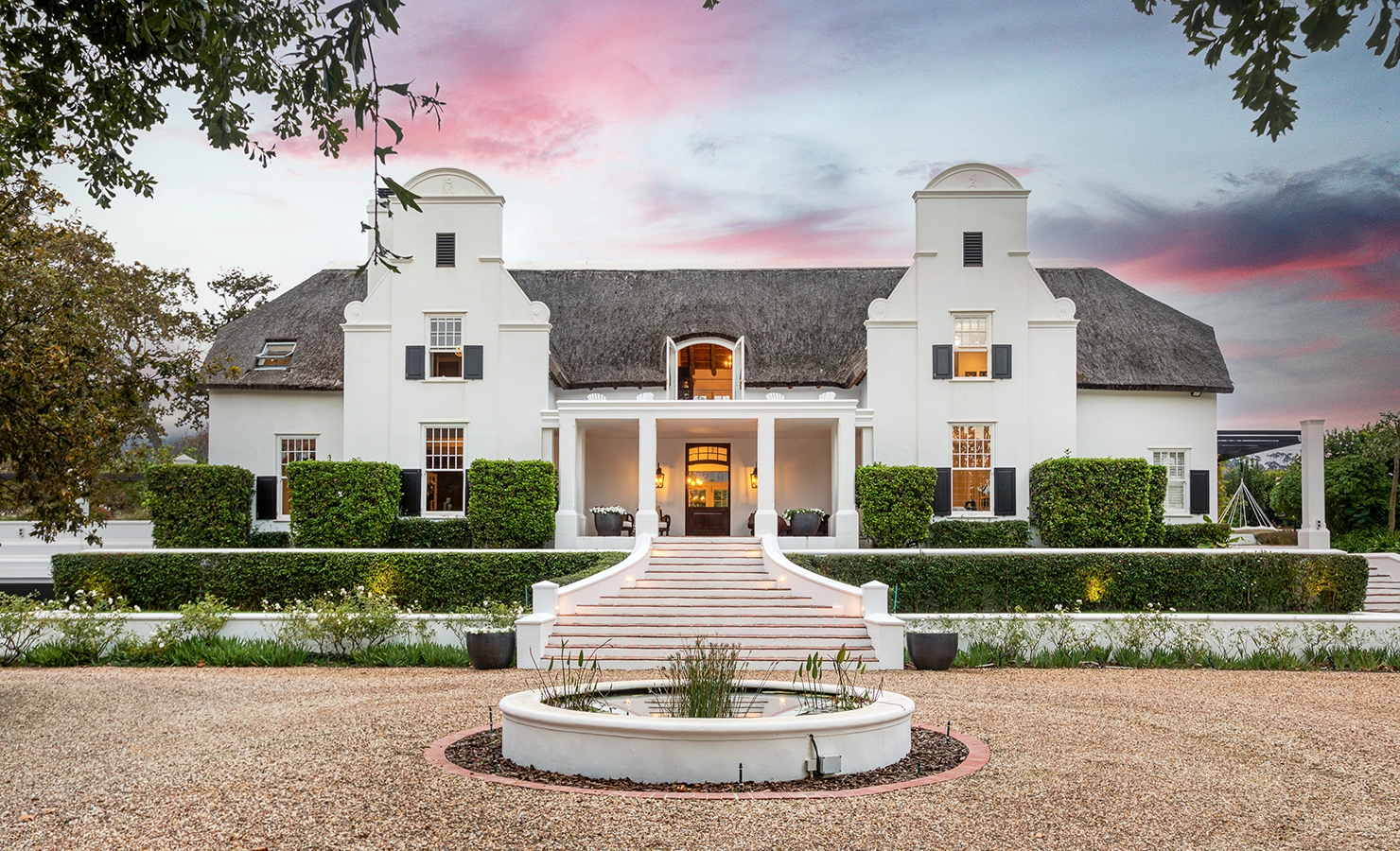 Perfect Hideaways: A grand Cape Dutch manor with a thatched roof, whitewashed façade, and elegant gabled towers, set against a dramatic pink-hued sunset. A sweeping staircase leads to the warmly lit entrance, flanked by manicured hedges and a circular water feature in the foreground. Buitenzorg Estate, Constantia.
