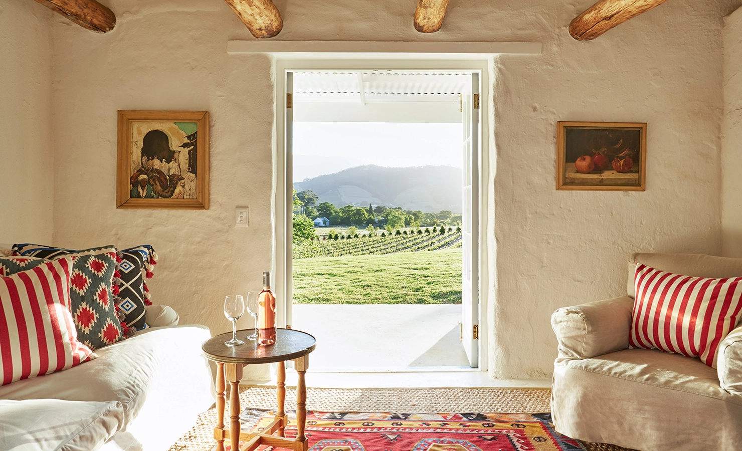 Perfect Hideaways: A rustic lounge with exposed wooden beams, patterned cushions on a sofa, and an open doorway leading to a view of vineyards bathed in golden light. La Cotte Forest Cottages, Franschhoek.
