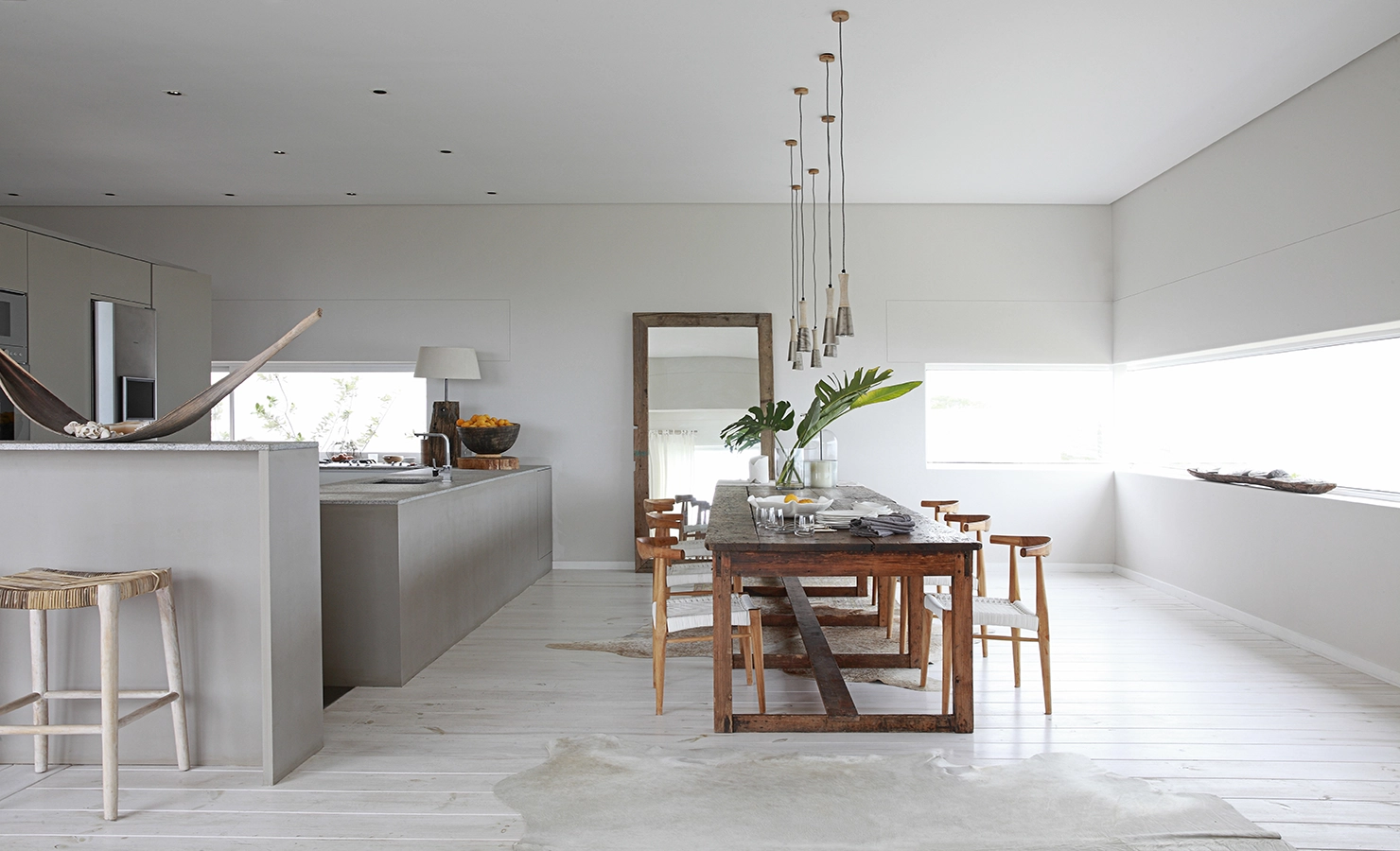 Perfect Hideaways: A serene open-plan kitchen and dining area with a minimalist coastal aesthetic, featuring a rustic wooden dining table, woven chairs, and pendant lighting. Soft neutral tones, natural textures, and large windows invite an abundance of light, creating a tranquil atmosphere. Salt House, Yzerfontein.