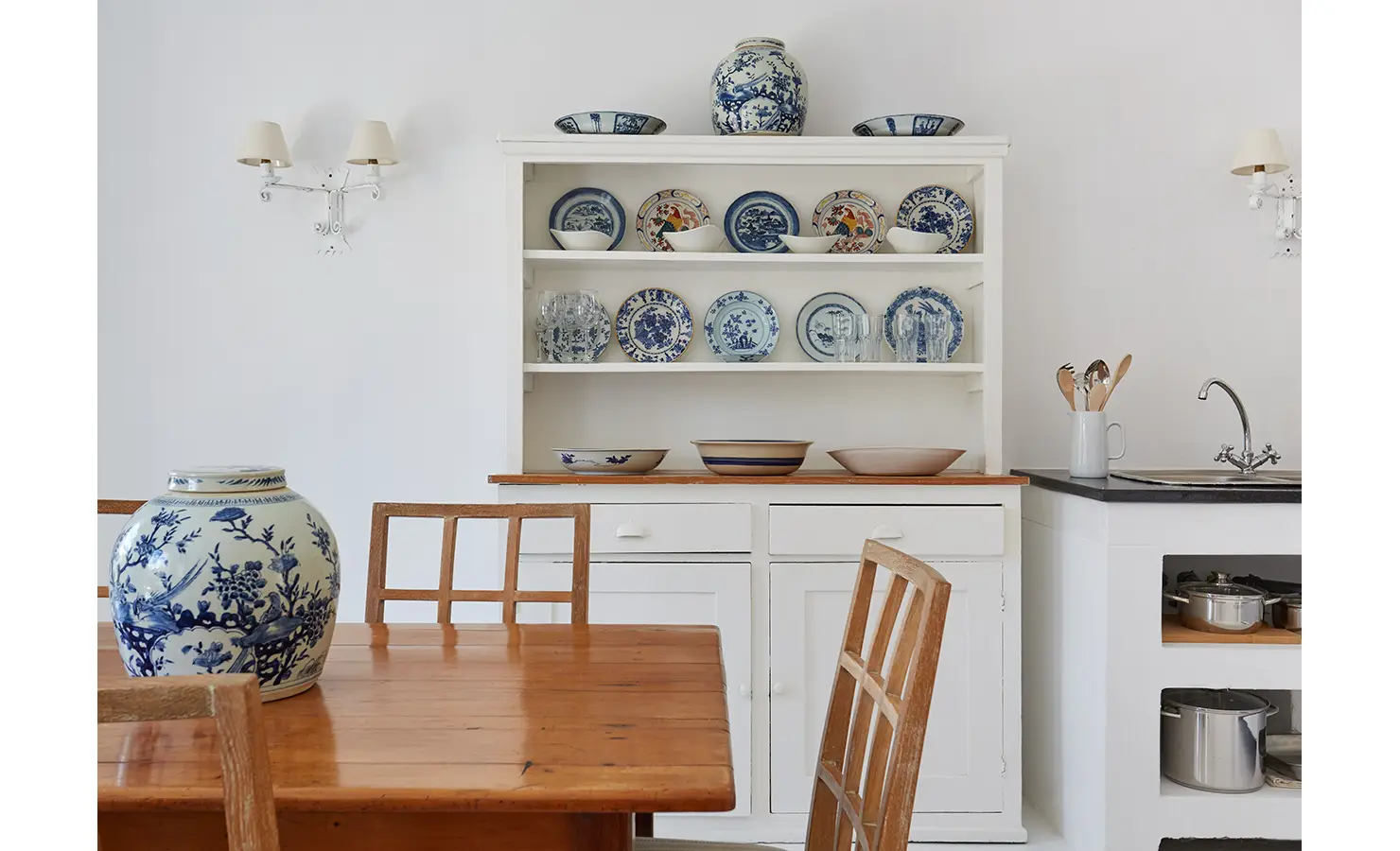 Perfect Hideaways: A cosy kitchen with a wooden dining table, blue-and-white ceramics on display, and a sink area, all set against white walls. La Cotte Forest Cottages, Franschhoek.