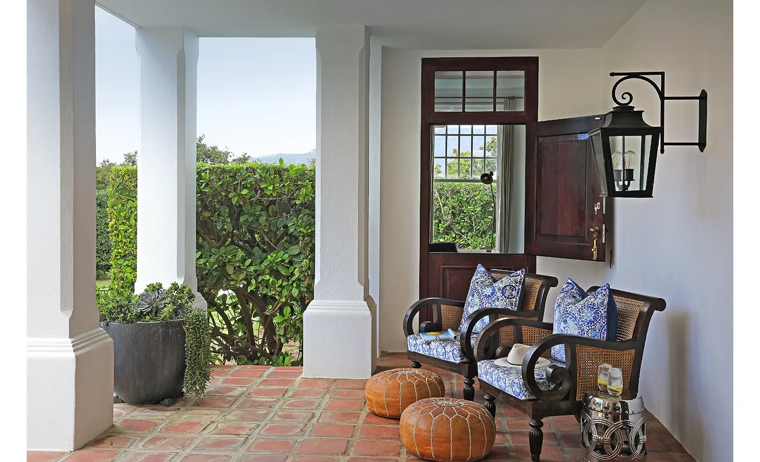 Perfect Hideaways: A charming covered veranda with elegant white pillars and a rustic brick floor, offering a cosy seating area with cane chairs adorned with blue patterned cushions. A wooden stable door opens to lush greenery. Buitenzorg Estate, Constantia.