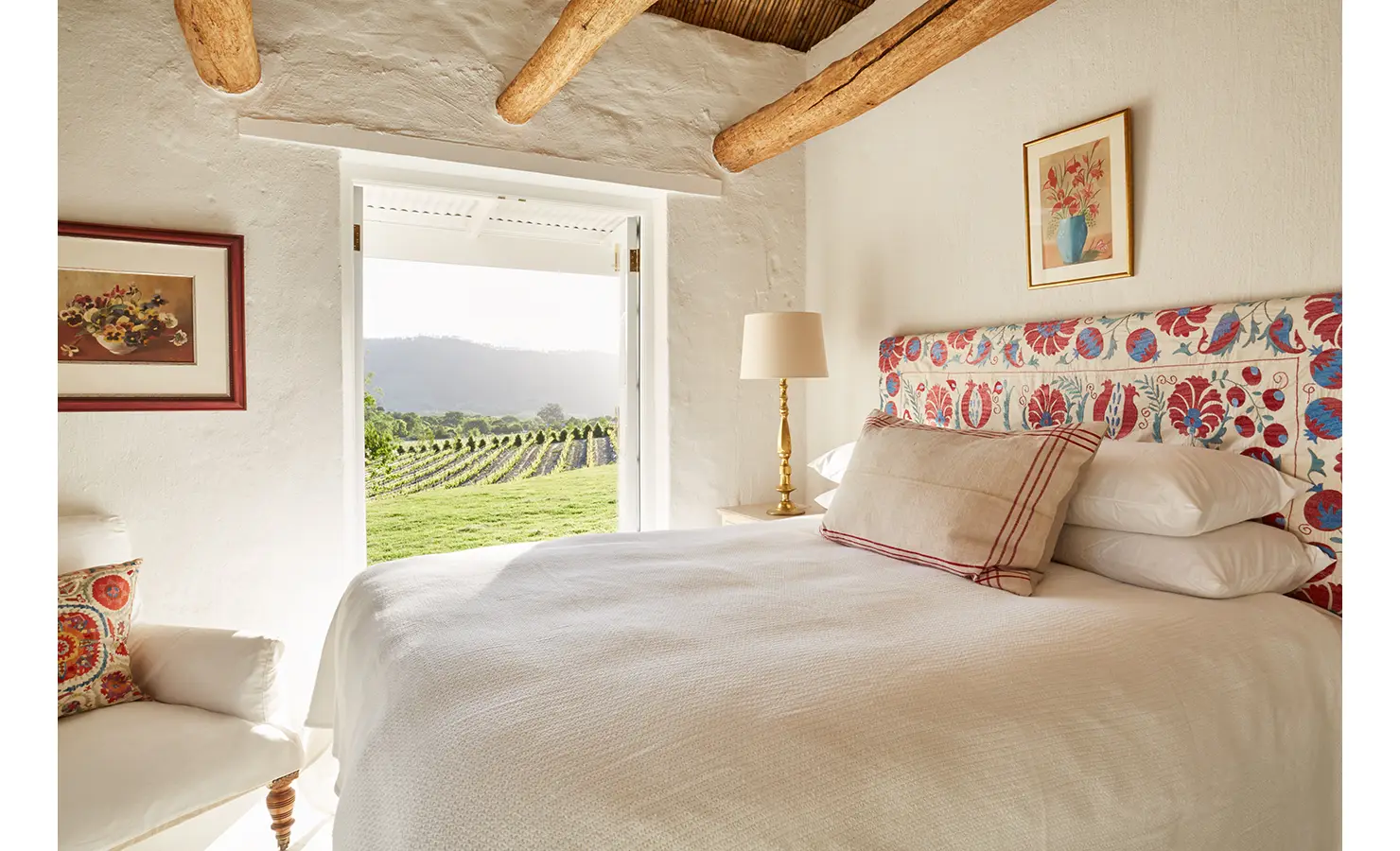 Perfect Hideaways: A charming bedroom featuring a colourful floral headboard, crisp white bedding, and an open doorway offering views of lush vineyards. La Cotte Forest Cottages, Franschhoek.