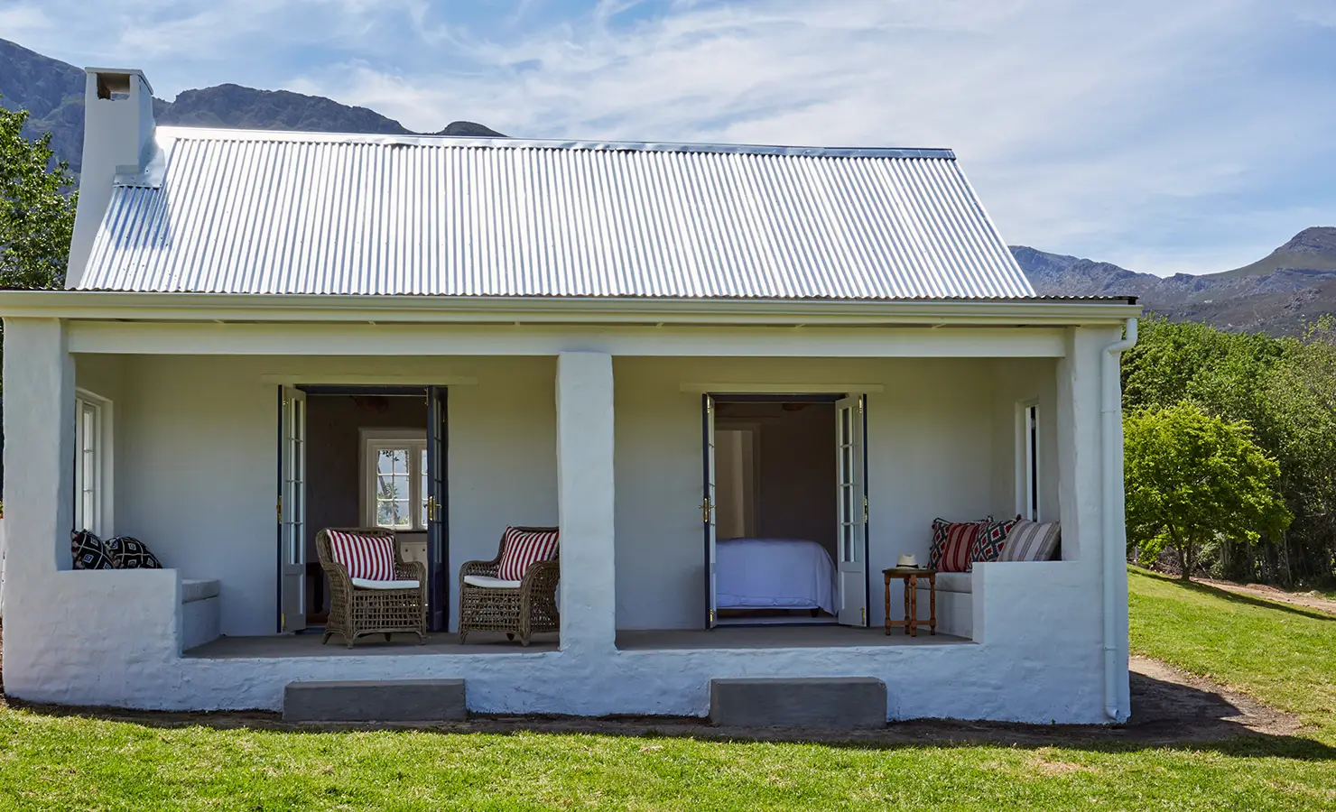 Perfect Hideaways: A quaint white cottage with a tin roof, featuring two open verandas furnished with wicker chairs and striped cushions, surrounded by greenery and mountain views. La Cotte Forest Cottages, Franschhoek.