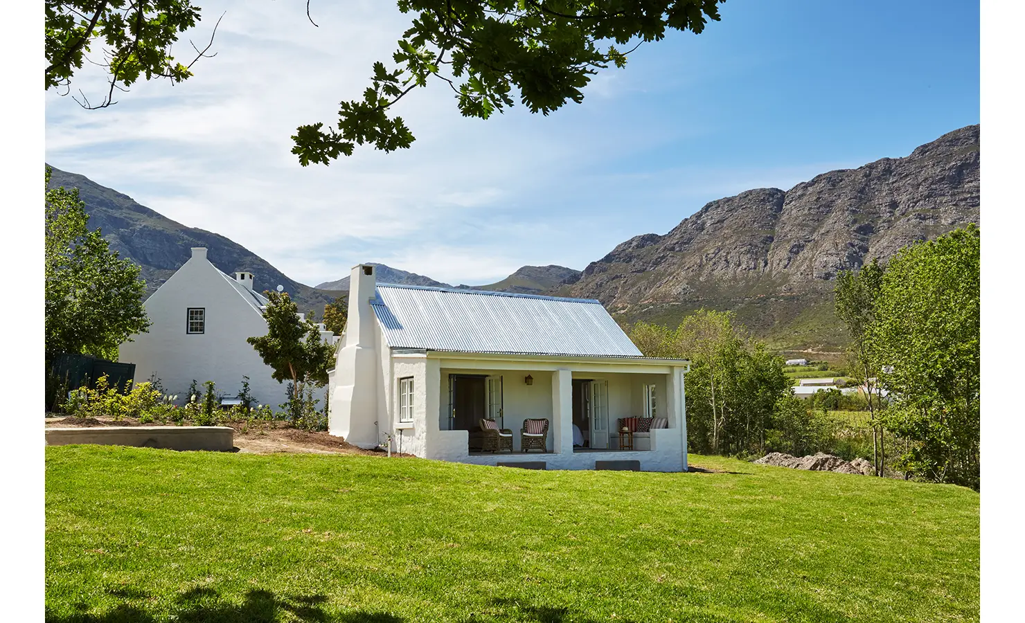 Perfect Hideaways: An exterior view of a traditional white Cape Dutch-style cottage, surrounded by a lush lawn and framed by dramatic mountain backdrops. La Cotte Forest Cottages, Franschhoek.