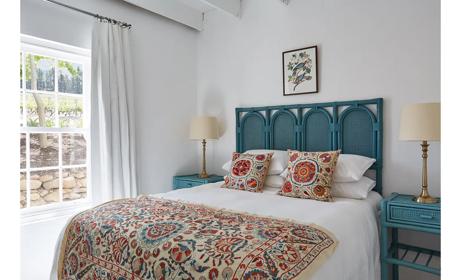 Perfect Hideaways: A light-filled bedroom with a turquoise headboard, colourful patterned bedding, and large windows overlooking a serene vineyard. La Cotte Forest Cottages, Franschhoek.