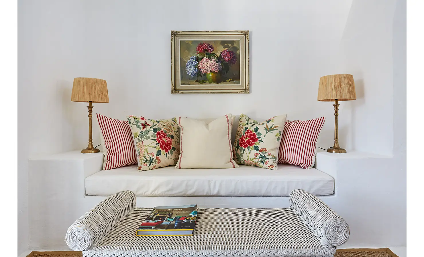Perfect Hideaways: A cosy built-in white sofa decorated with floral and striped cushions, flanked by brass table lamps, with a framed painting of hydrangeas above. La Cotte Forest Cottages, Franschhoek.
