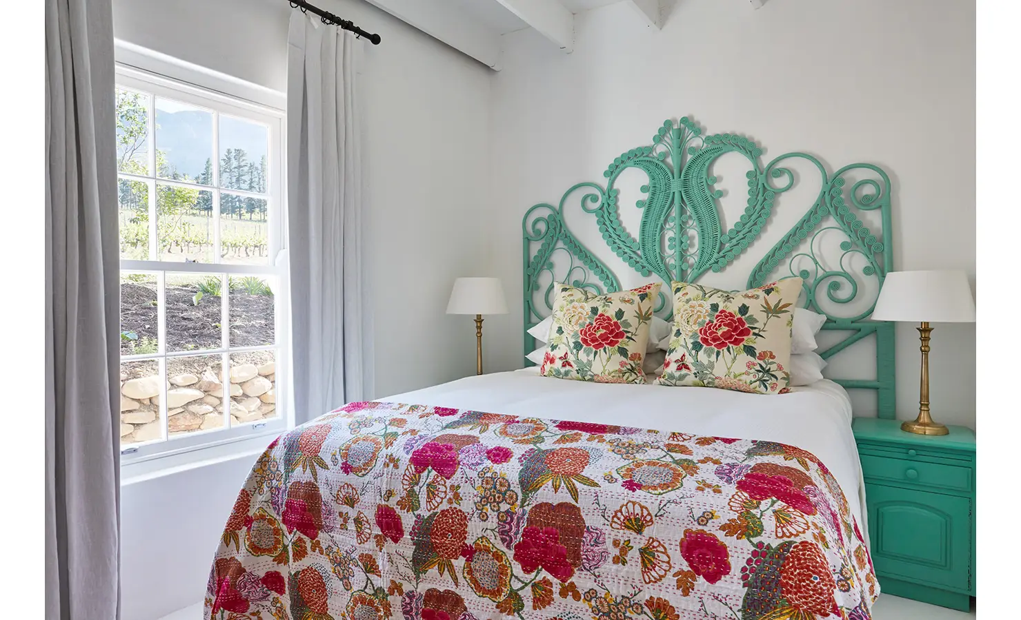 Perfect Hideaways: A light-filled bedroom featuring a turquoise ornate headboard, floral-patterned cushions, and a colourful quilt, with a view of vineyards through the window. La Cotte Forest Cottages, Franschhoek.